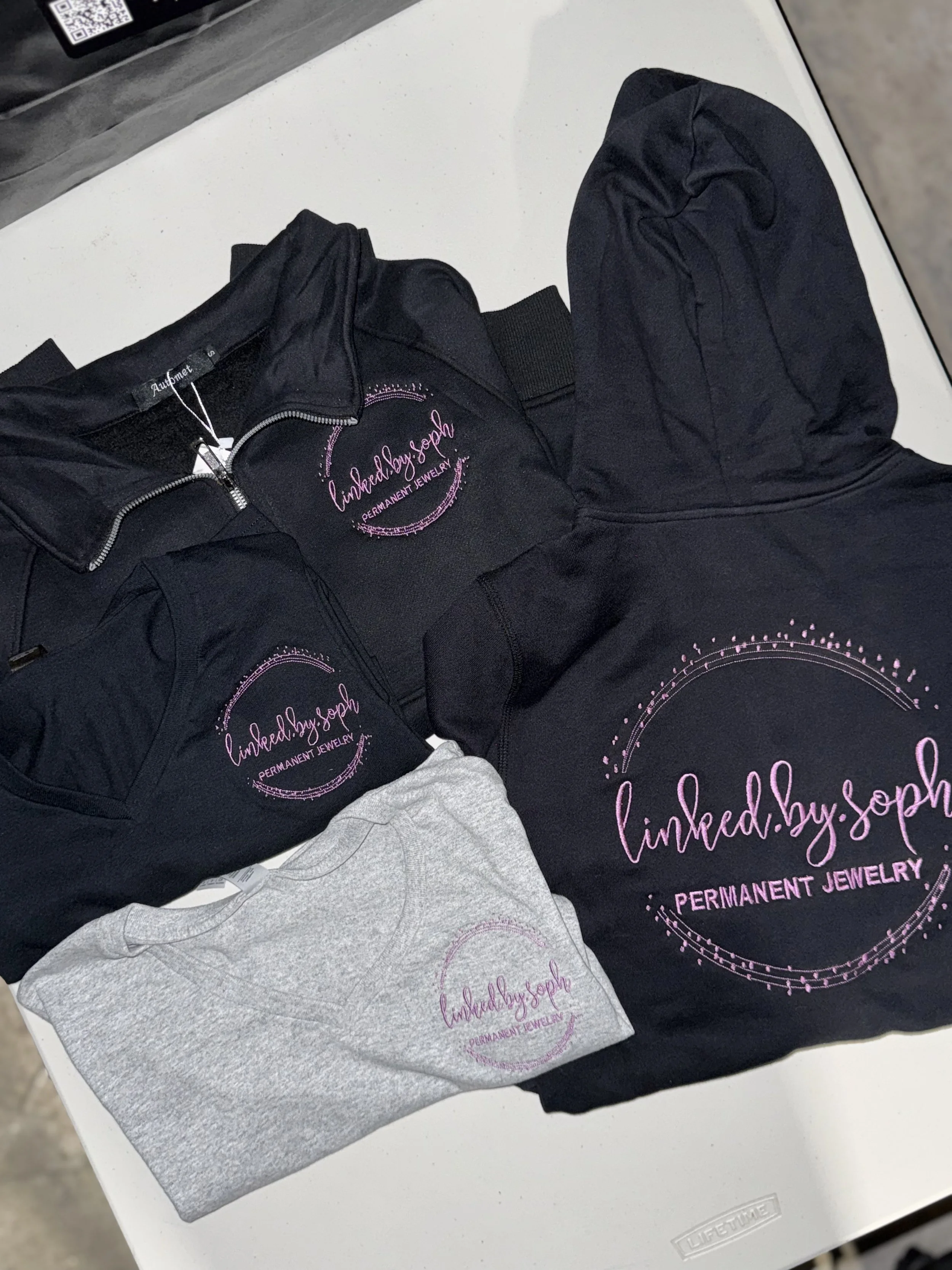 Clothing and accessories with 'linked.by.soph' logo, including black and gray shirts with pink embroidered text, displayed on a white table.