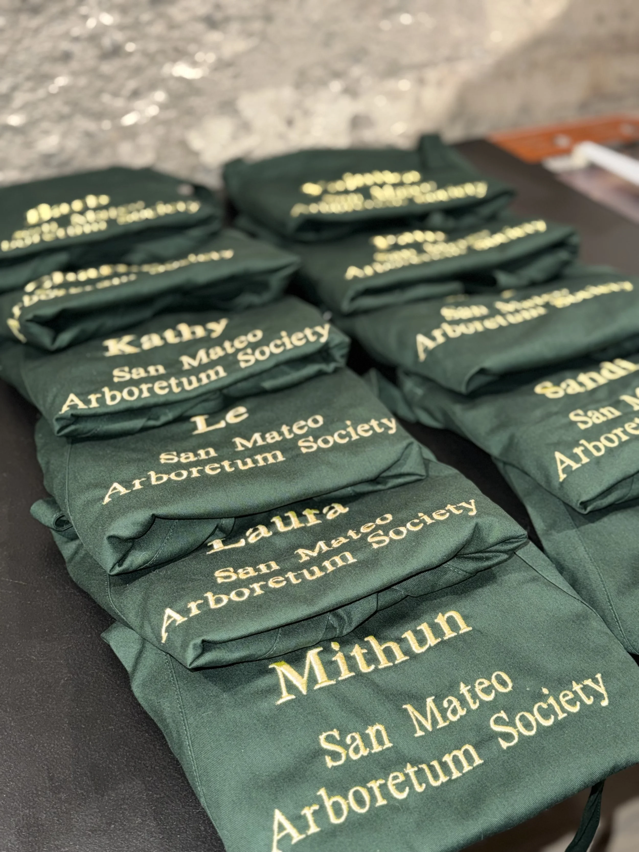 Green fabric bags with yellow text that read 'Mithun, San Mateo Arboretum Society', stacked on a dark table.