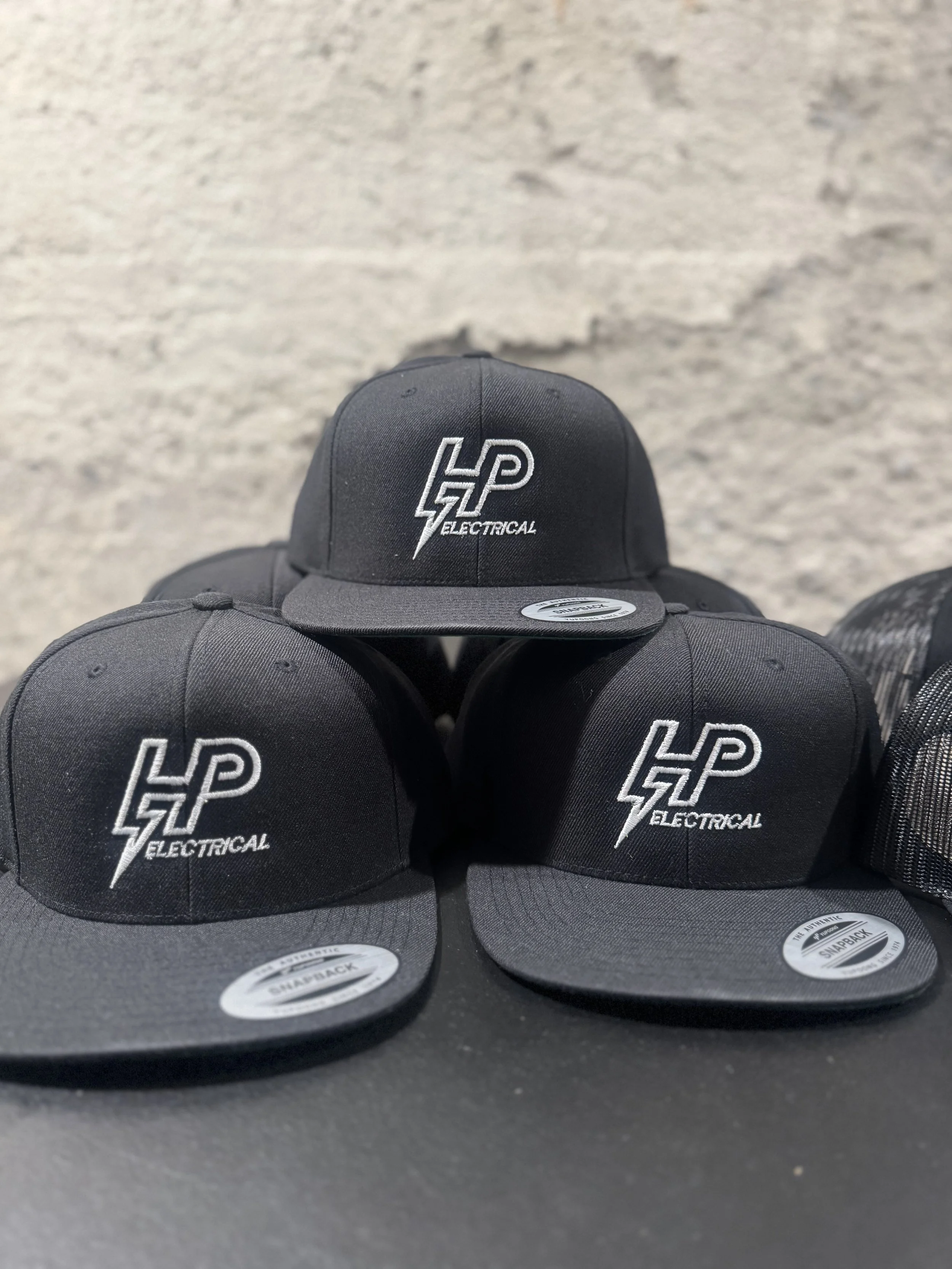 Three black baseball caps with white embroidered logo and text that reads HP Electrical, displayed on a black surface against a gray textured background.