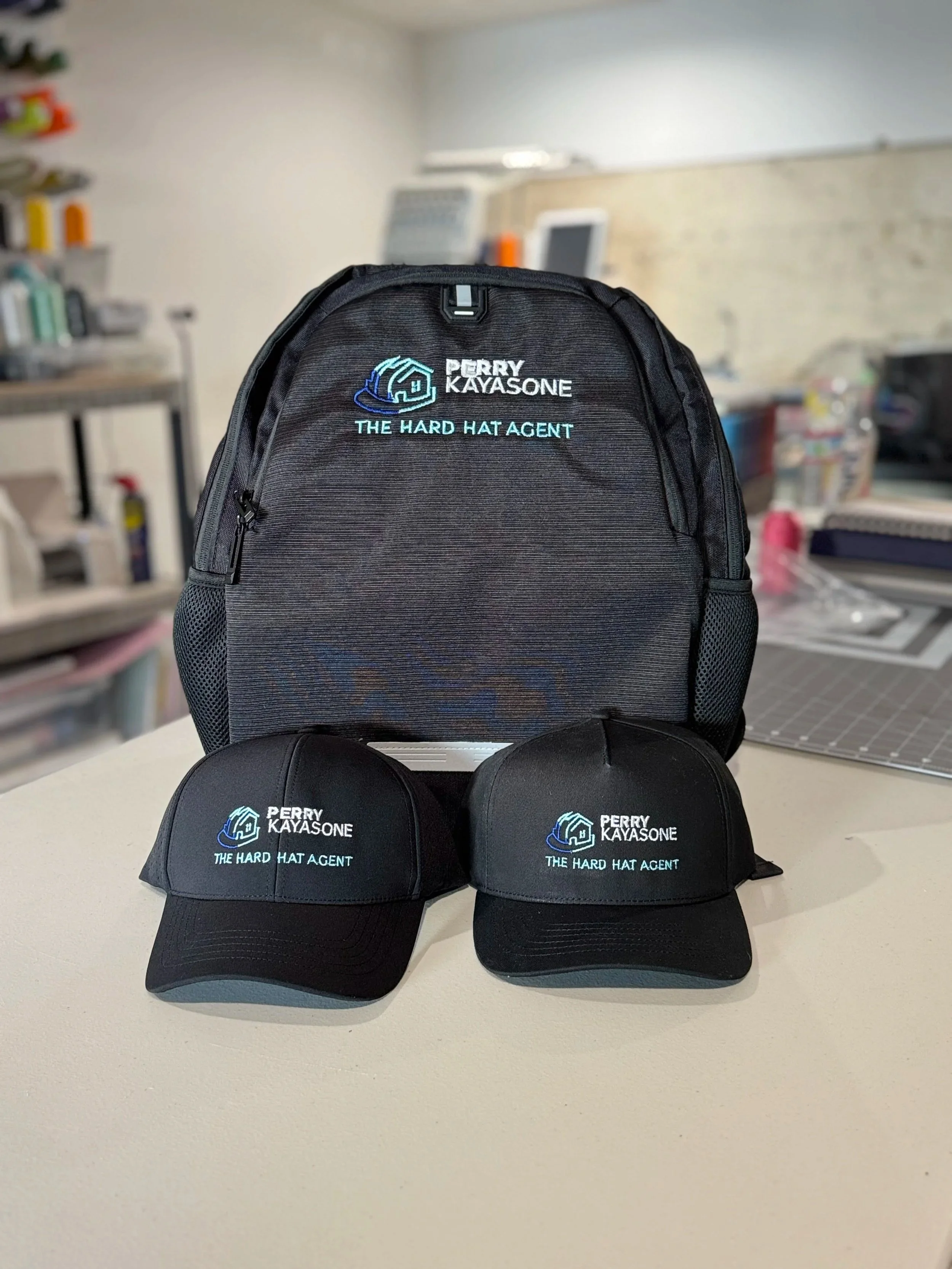 Set of black cap and backpack with embroidered logo and text reading 'Perry Kayasone, The Hard Hat Agent' on a white surface.