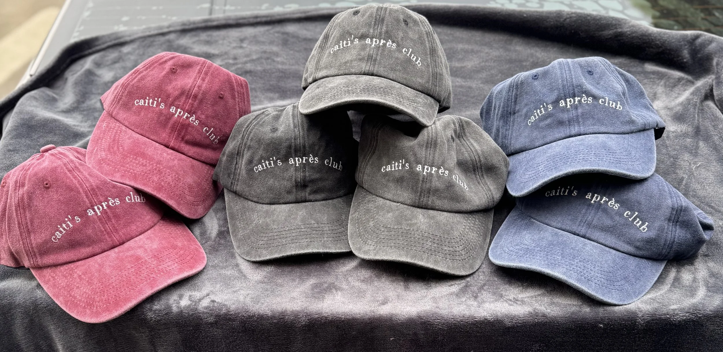 Six corduroy baseball caps in pink, gray, and blue colors with embroidered text 'caiti's après club' on the front, arranged on a gray fabric surface.