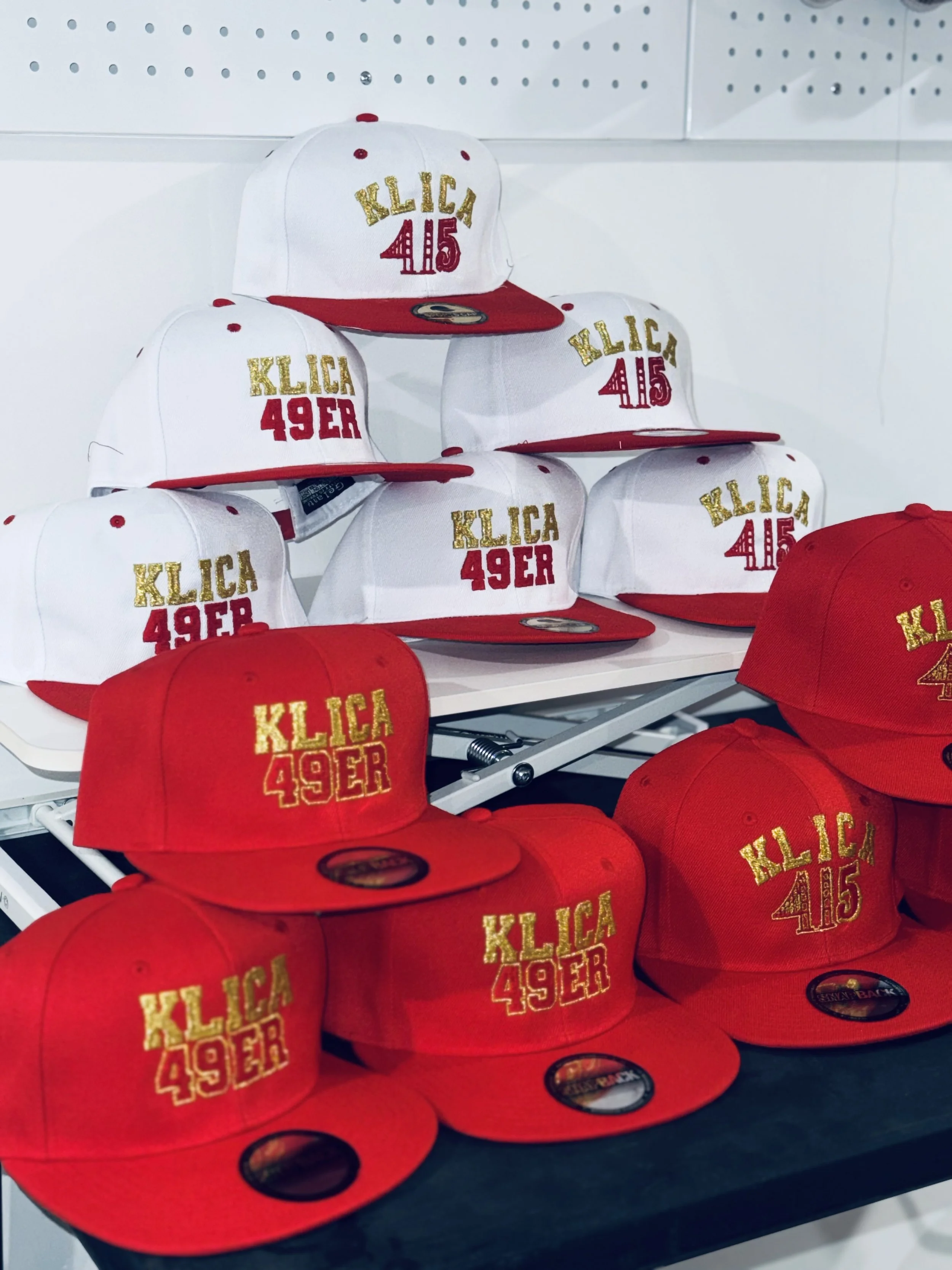 Display of red and white baseball caps with gold and red embroidery reading "KLICA 49ER" and "KLICA 415" on shelves in a retail store.