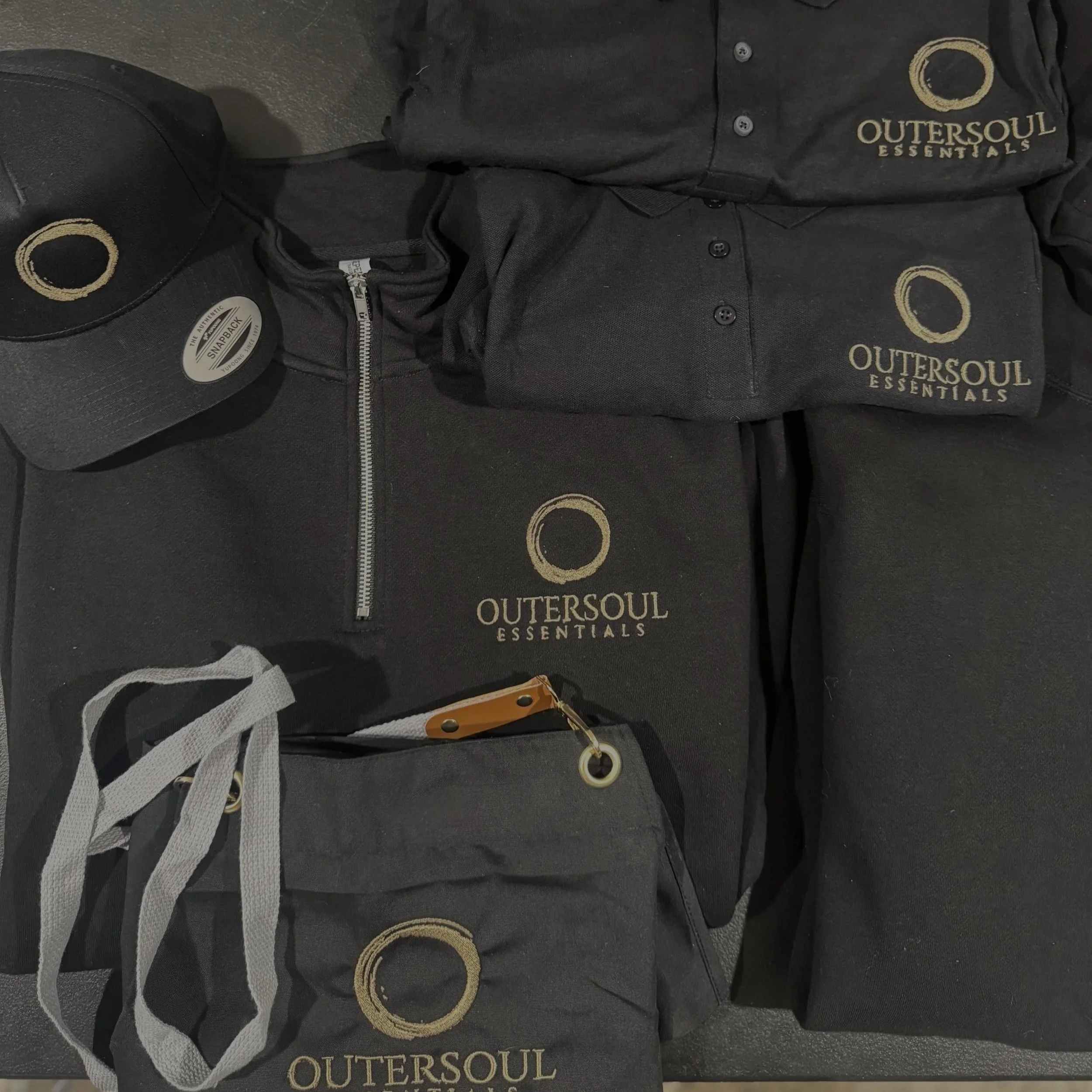 Various embroidered black clothing and accessories branded with Outersoul Essentials logo, including a cap, t-shirts, a polo shirt, a hoodie, a drawstring bag, and a small pouch.