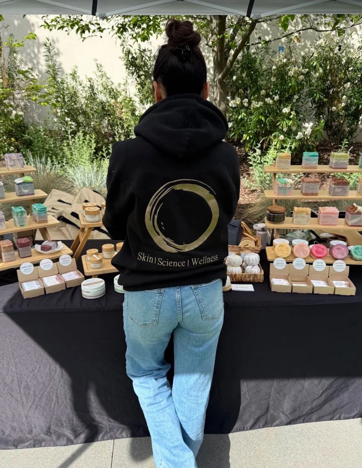Person standing at a booth with embroidered logo on sweatshirt selling skincare and wellness products outdoors, with display shelves of colorful containers and jars, under a canopy with trees and greenery in the background.