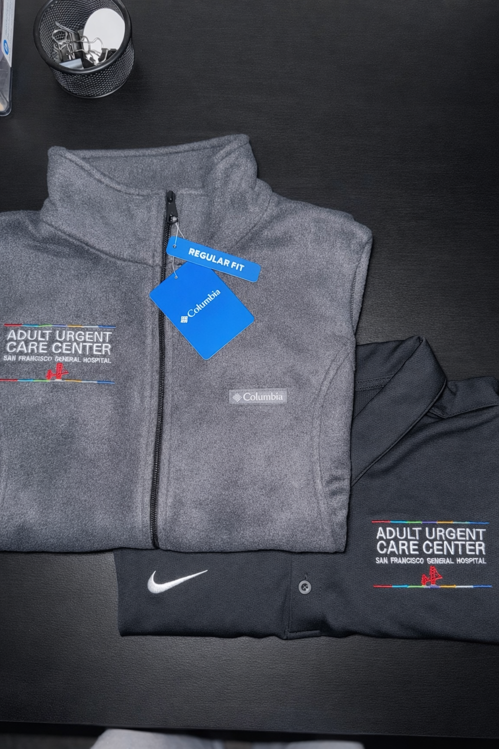 Two folded jackets, one gray Columbia fleece with a zip and a tag that reads 'Regular Fit,' and the other black with a white Nike logo, are placed on a black surface. There is also a small black container with paper clips and a white object in the to