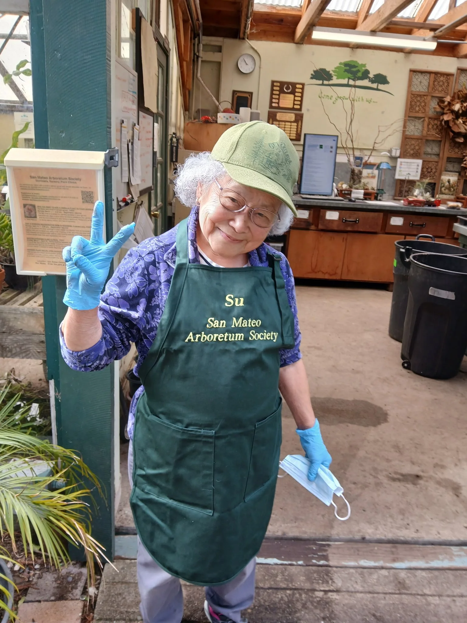 An elderly woman with gray curly hair, wearing glasses, a green cap, a purple blouse, and a green apron that reads "Su San Mateo Arboretum Society." She is smiling and making a peace sign with her right hand while holding a face mask in her left hand