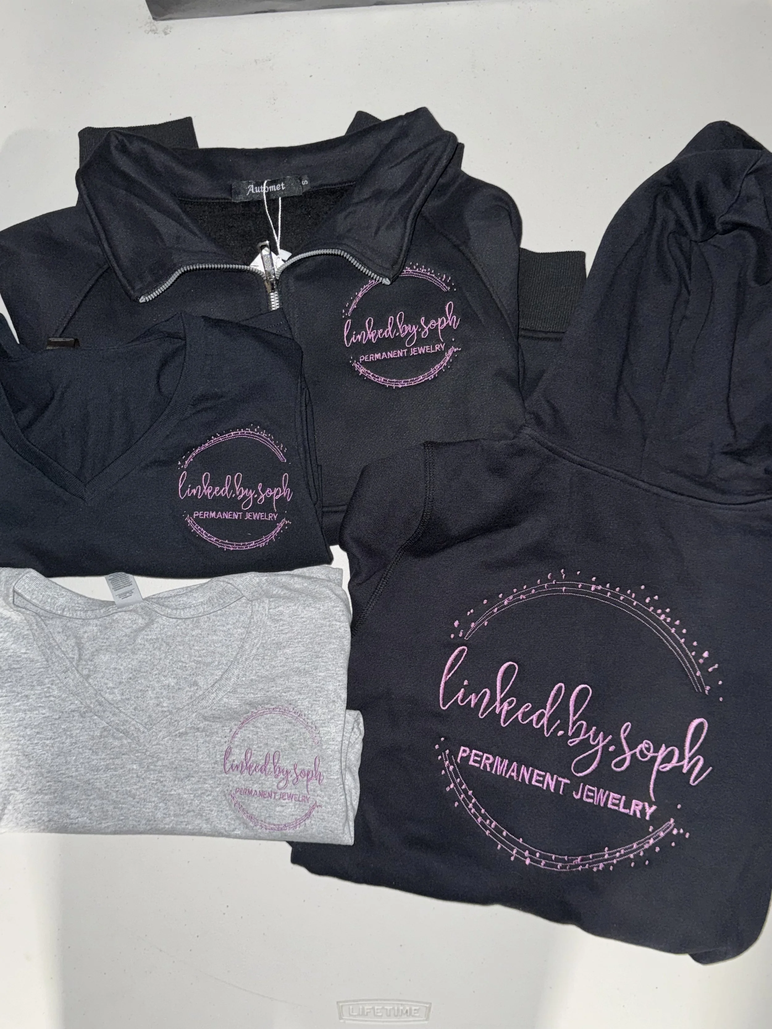 A collection of clothing items, including a black zip-up jacket, a black hoodie, and a gray t-shirt, all featuring pink embroidered logos of 'linked.by.soph' with the subtitle 'PERMANENT JEWELRY'.