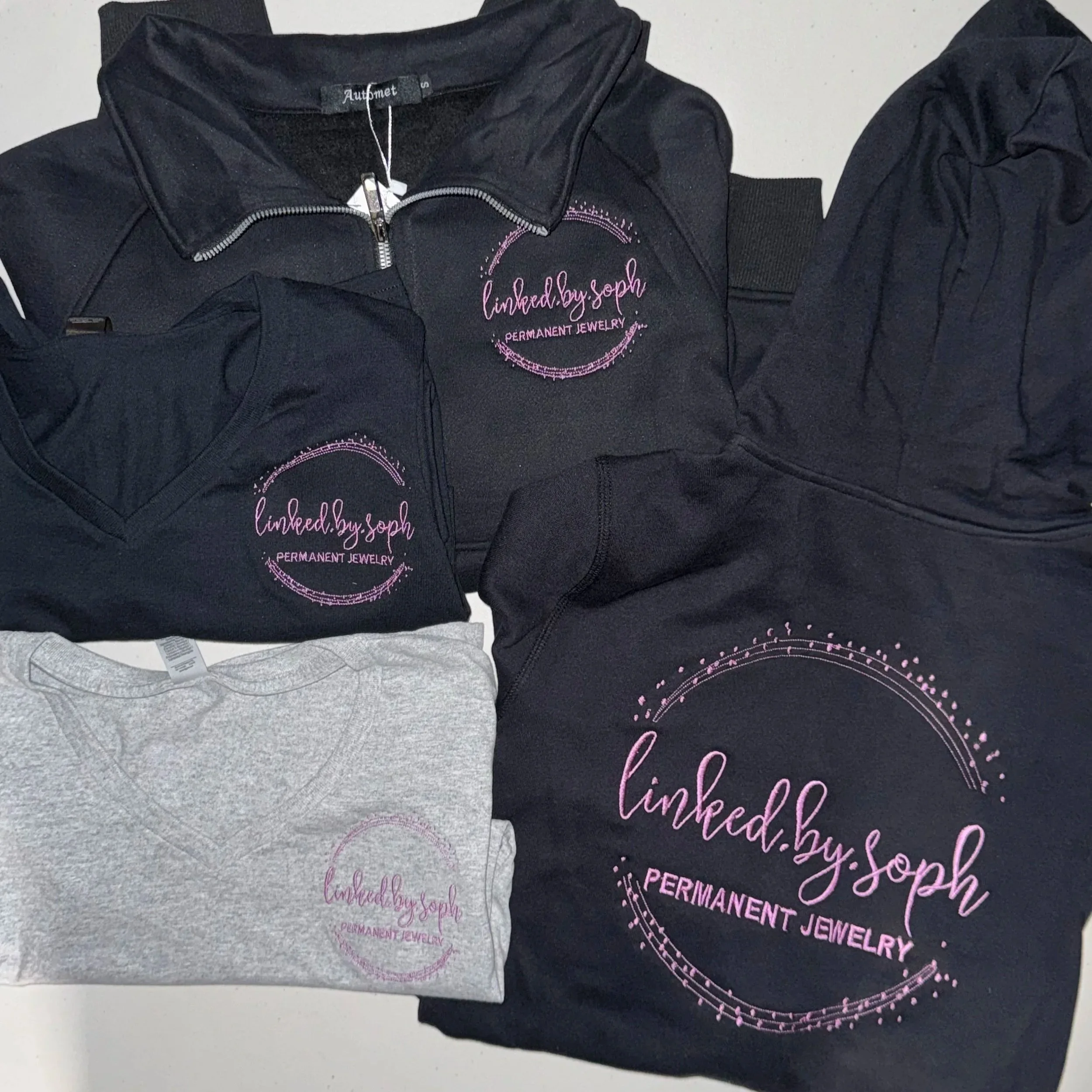 Various embroidered black and gray clothing items, including t-shirts, hoodies, and a bag, all with a pink logo that says 'linked.by.soph - PERMANENT JEWELRY' surrounded by decorative pink dots.