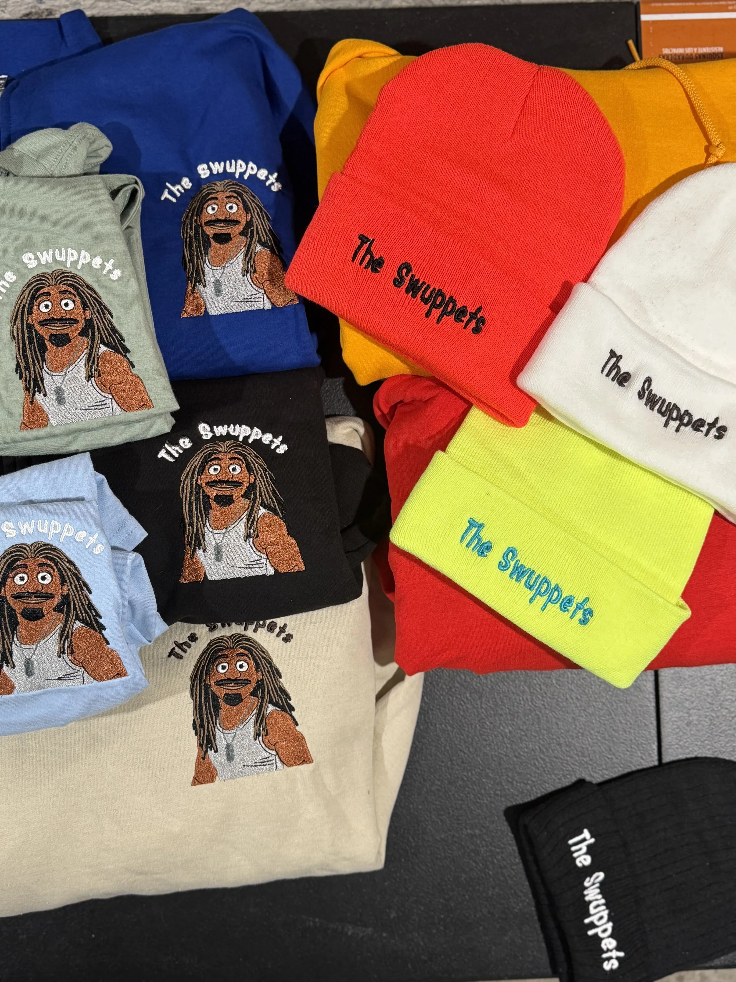 Various colorful beanies and t-shirts with a cartoon character and the text 'The Swuppets' displayed on a table.