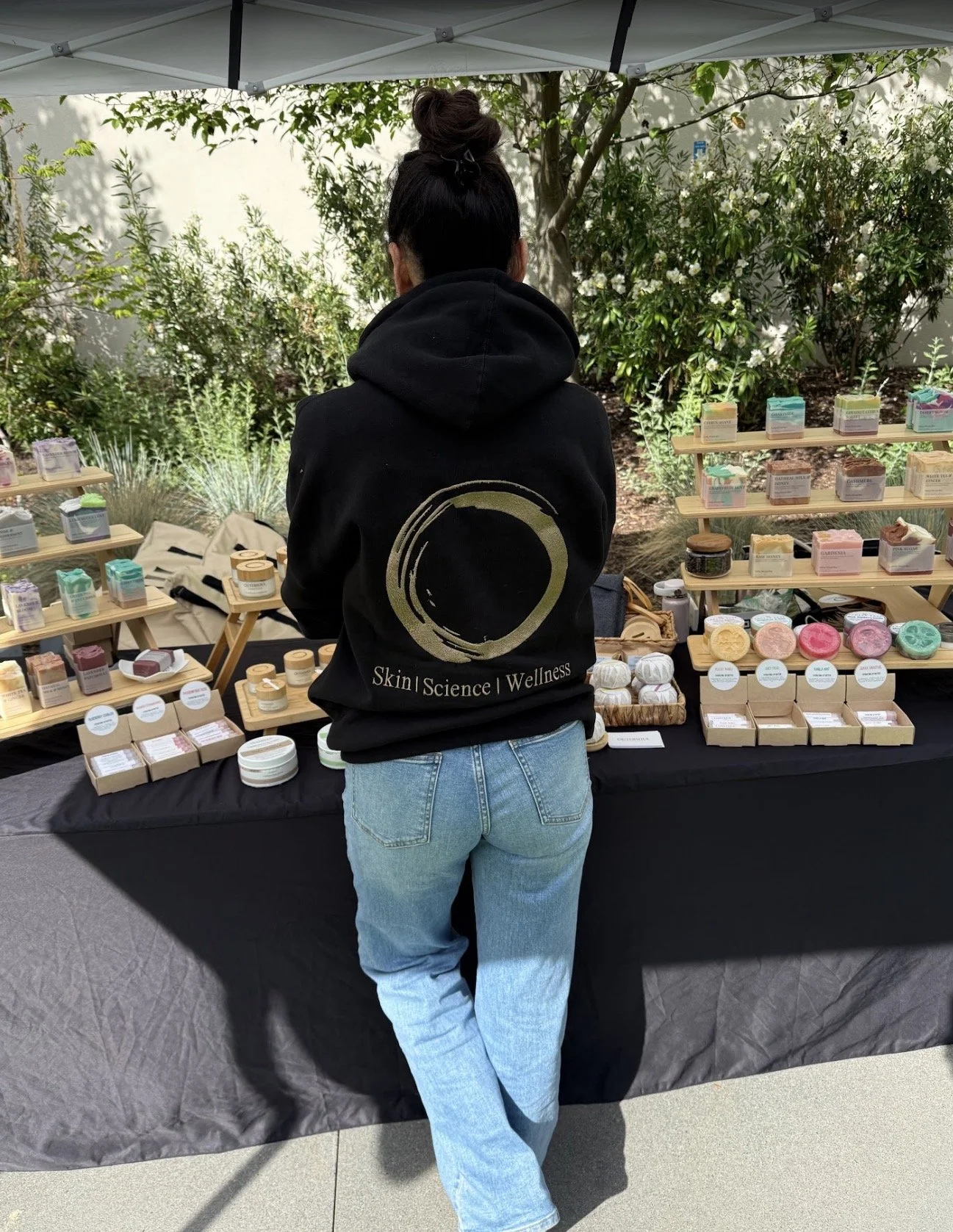 Person with a top knot hairstyle wearing a black hoodie that says "Skin | Science | Wellness" on the back, standing at a market stall selling skincare or soap products, with greenery and trees in the background.