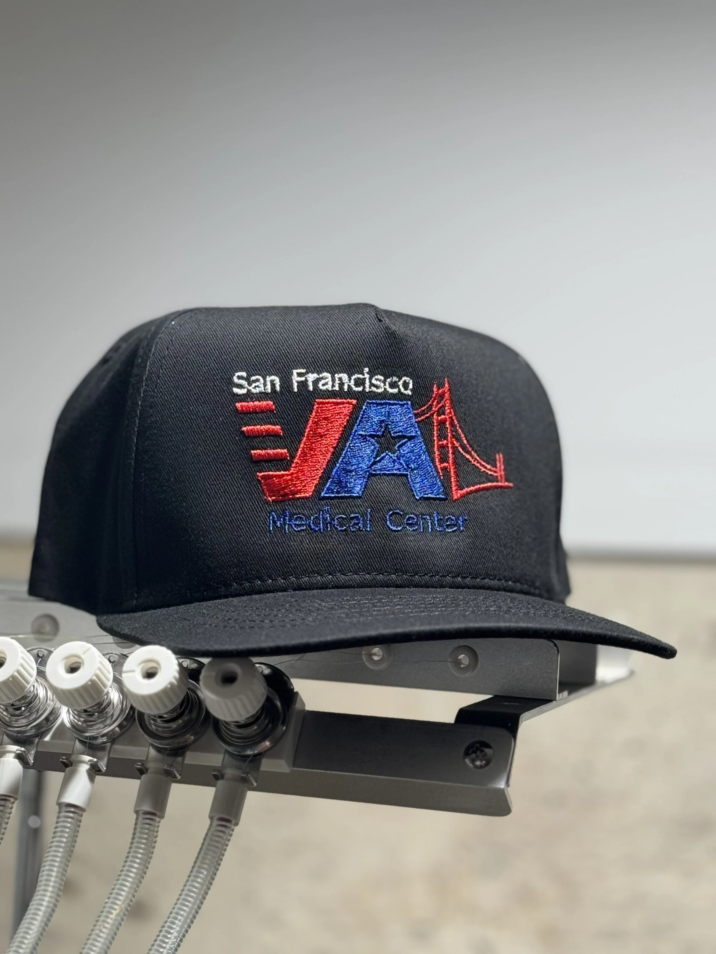 Black cap with embroidered logo for San Francisco VA Medical Center, featuring a red bridge, red and blue letters, and a star