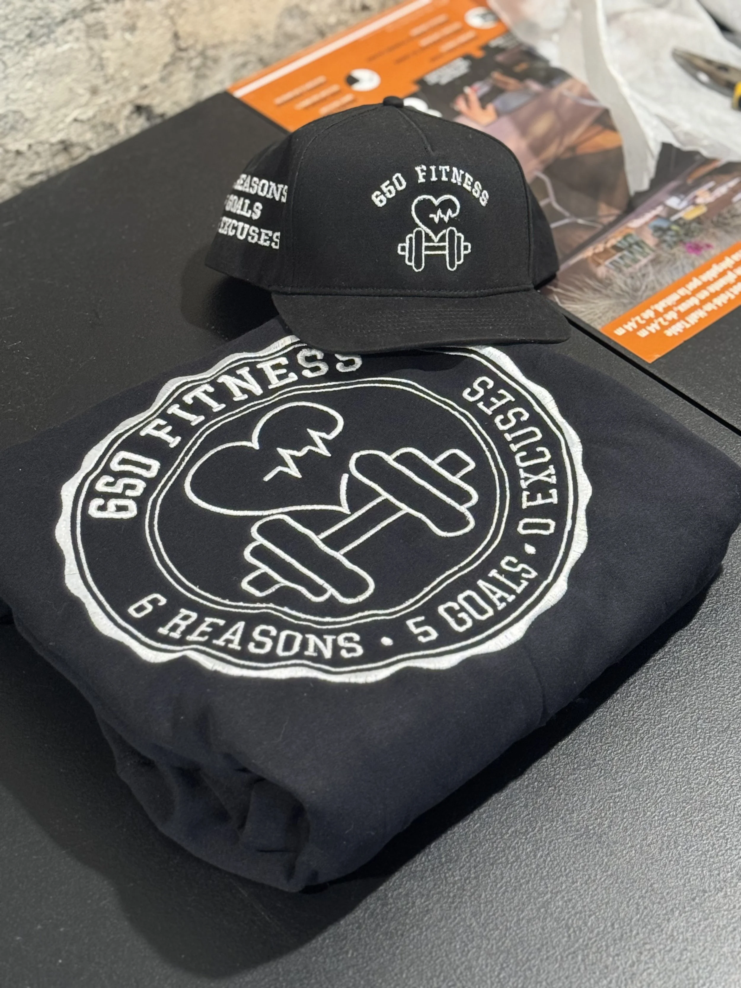 A black baseball cap and a black folded t-shirt with white designs and text are placed on a black surface, with an orange pamphlet and some paper in the background. The shirt features a logo with a heart, a dumbbell, and an electrocardiogram line, al