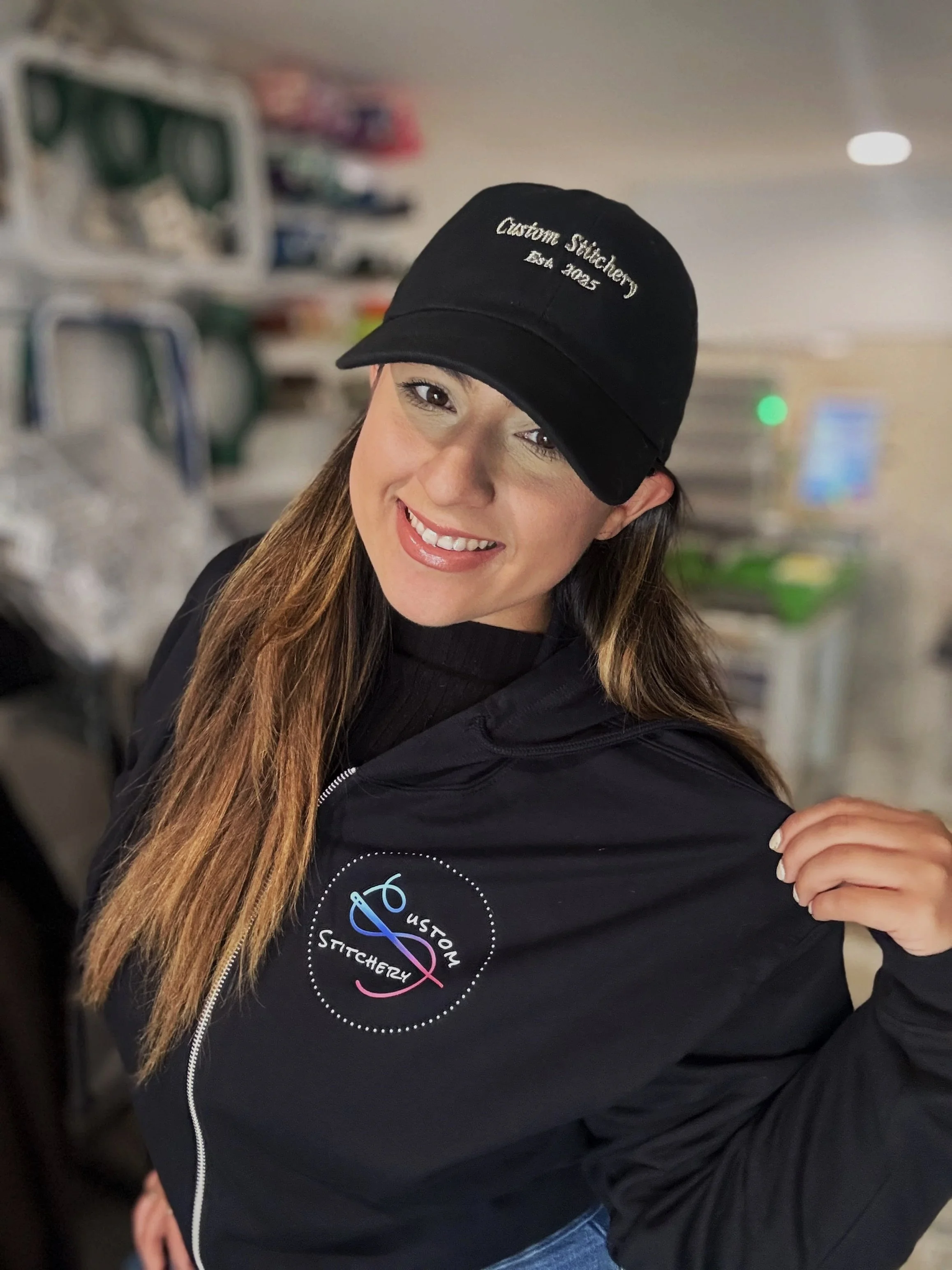 Smiling woman wearing a black cap and jacket with a custom stitchery logo.