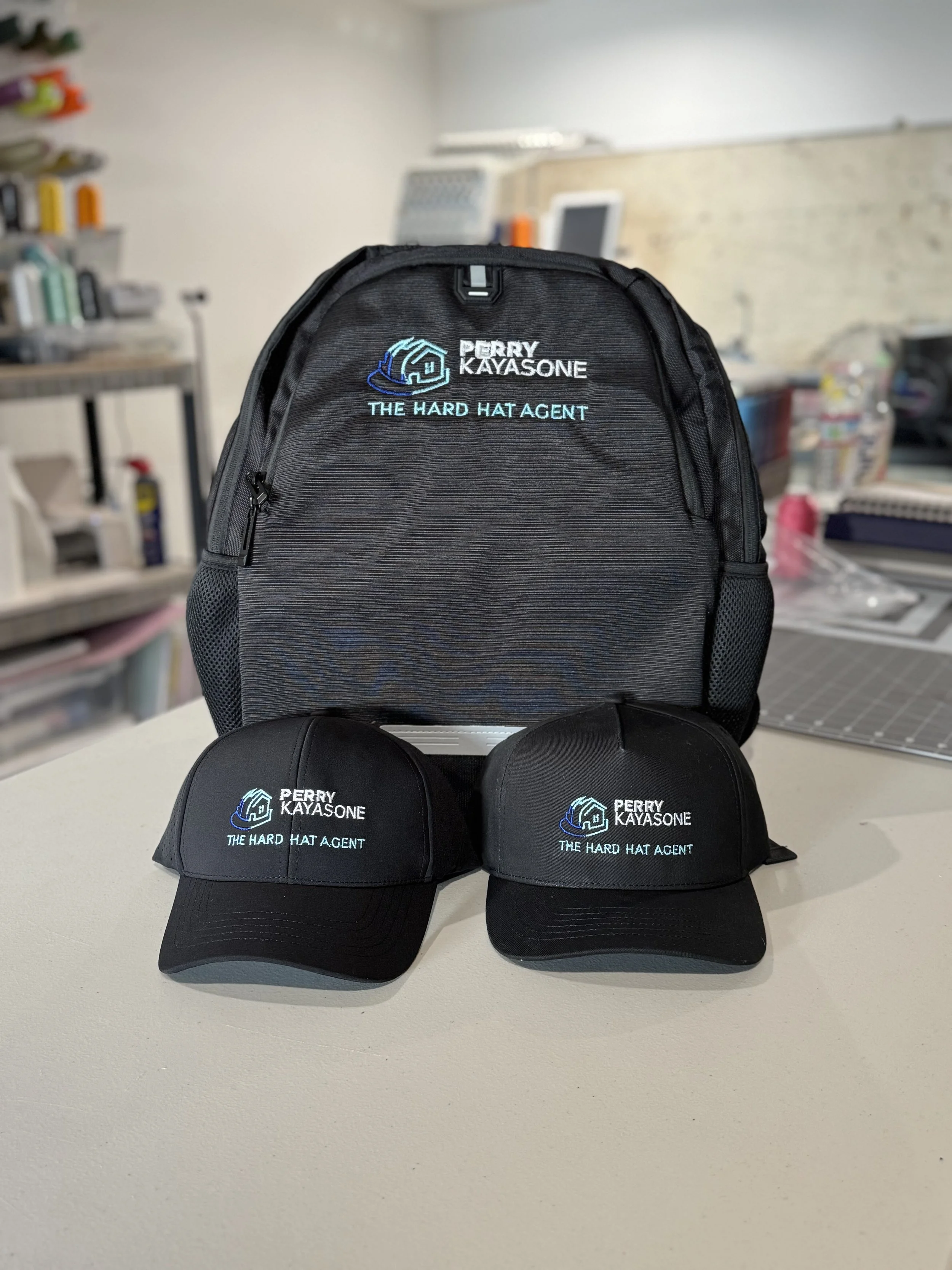 Black backpack and two black caps with embroidered logos and text, on a white table in a room.