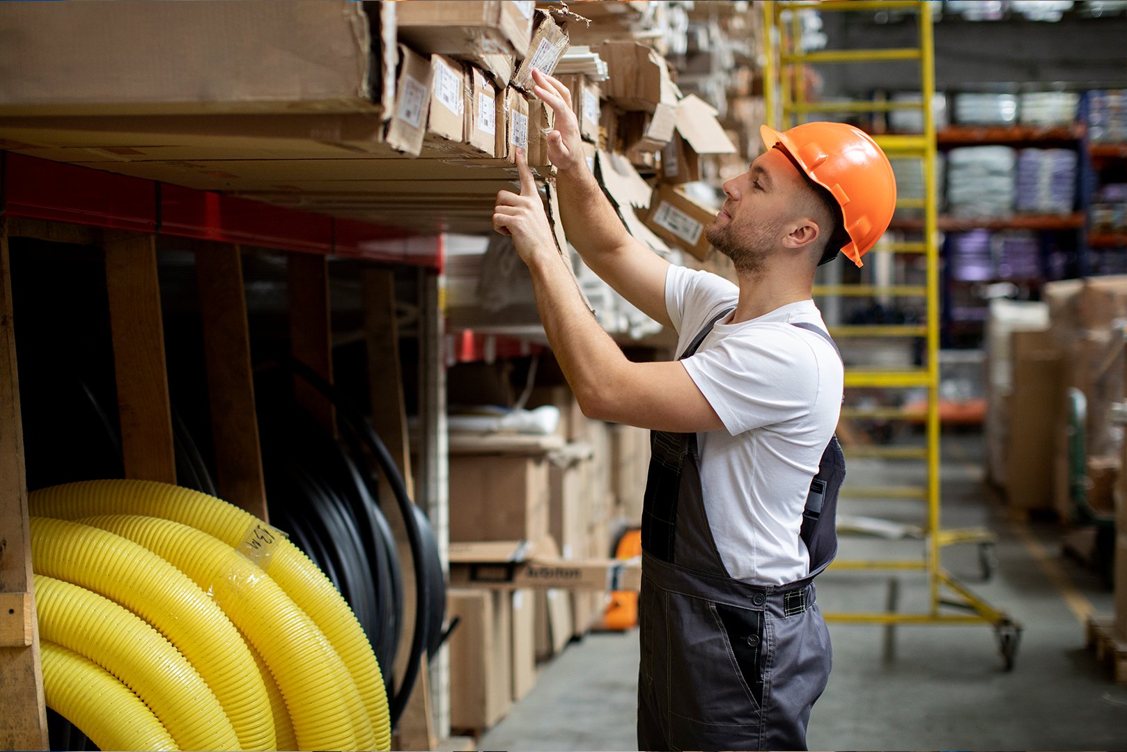 Hombre en un almacén inspeccionando productos en estantes, usando un casco de seguridad naranja.
