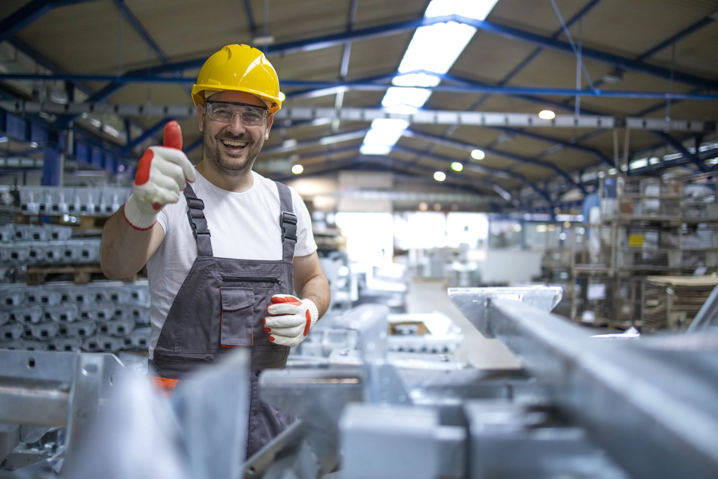 Hombre trabajador en una fábrica, usando casco de seguridad, guantes, gafas protectoras, camiseta blanca y overol gris, sonriendo y haciendo un gesto de aprobación con el pulgar en un ambiente industrial con estructuras metálicas y maquinaria.