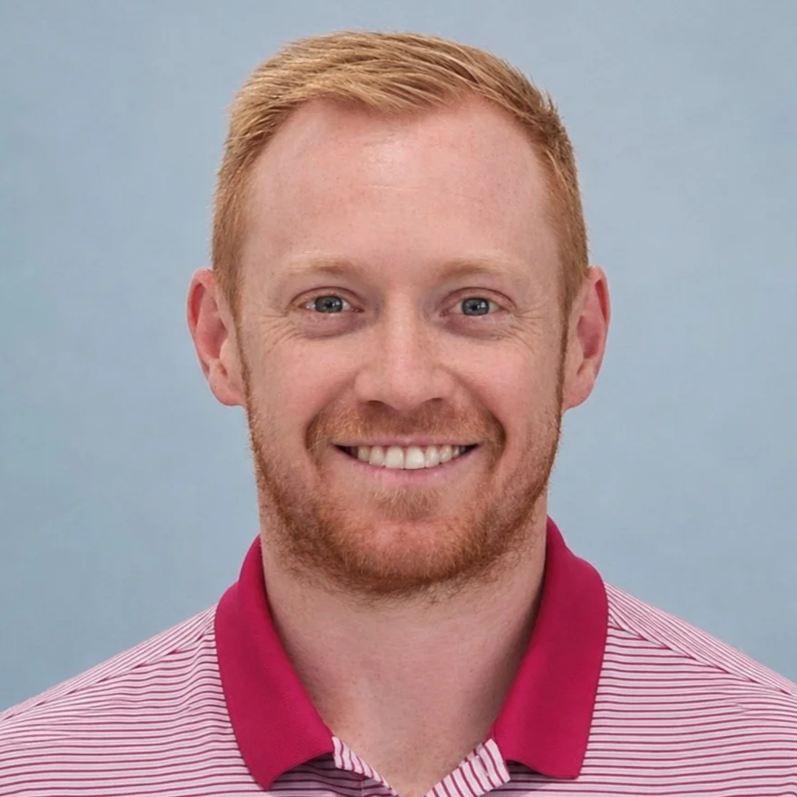 Close-up of a smiling man with red hair and beard wearing a red and white striped polo shirt.