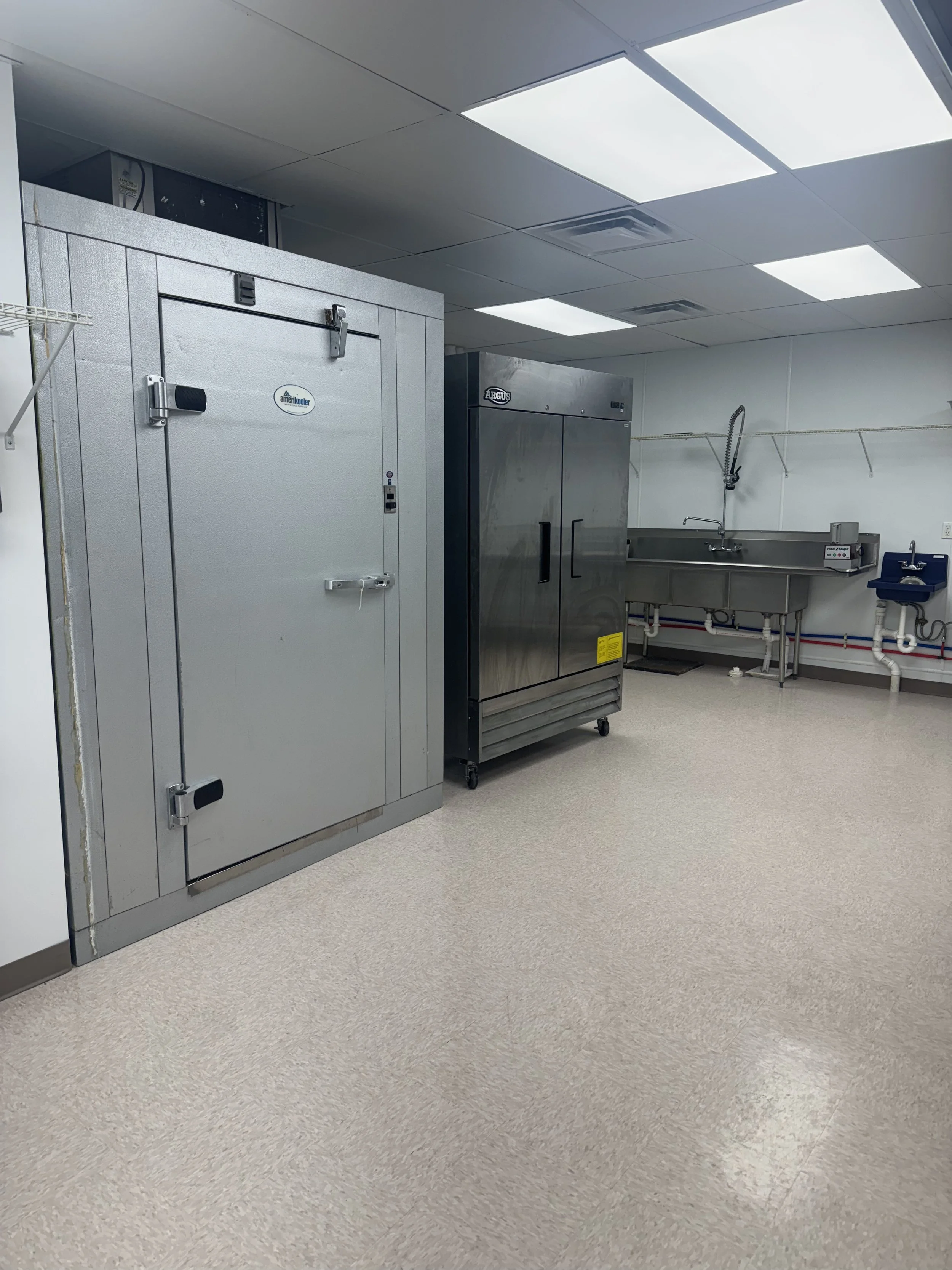 A commercial kitchen with a large gray walk-in freezer, a stainless steel refrigerator, and a stainless steel sink with a high-arc faucet. The room has white walls, a tiled floor, and ceiling lights.