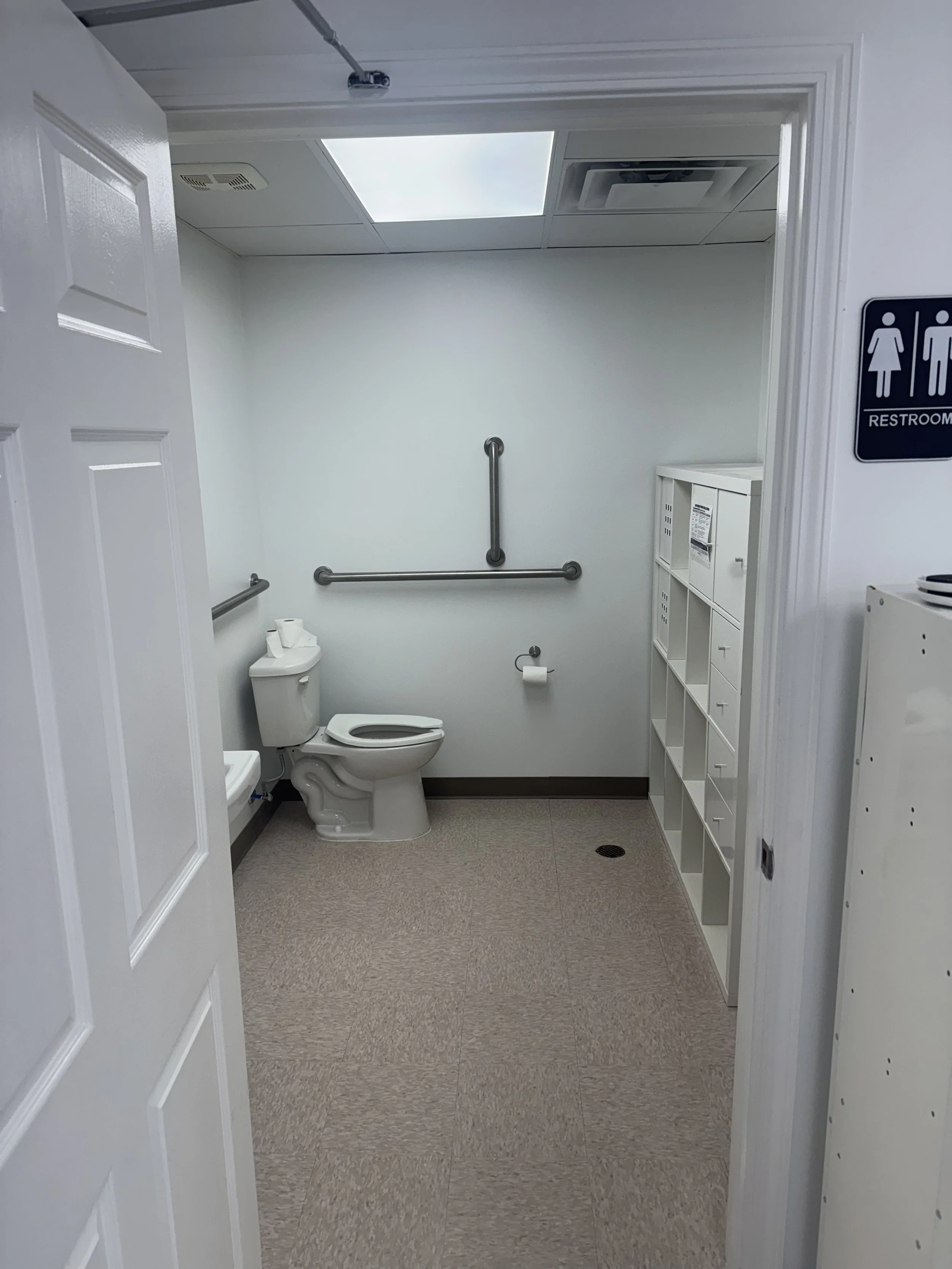Accessible restroom with white walls, beige tiled floor, and a ceiling with bright fluorescent lighting. Features include a toilet, a toilet paper holder, grab bars on the wall, and a white storage cubby unit. A sign indicating it is a women's restroom is visible outside the door.