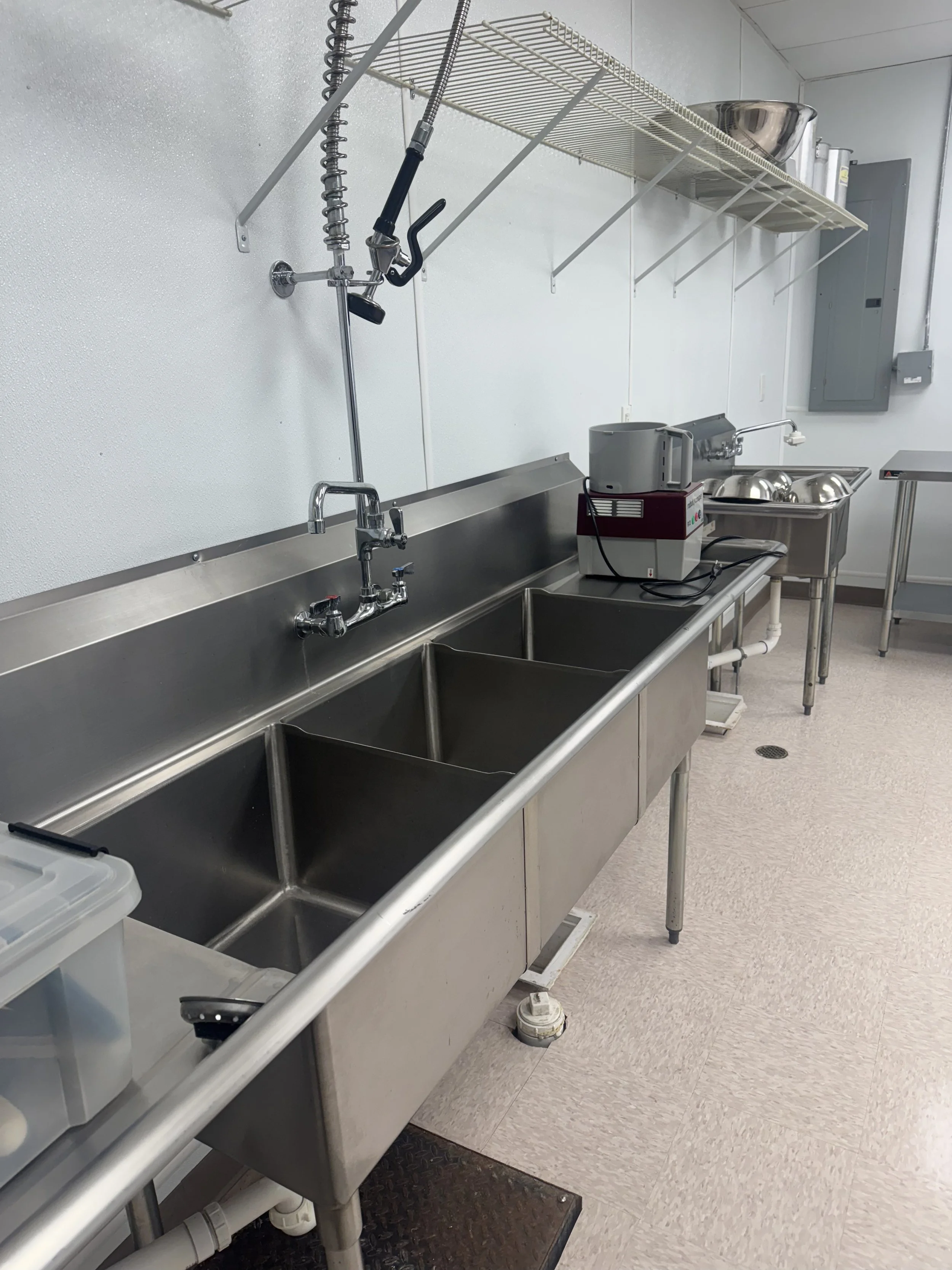 Commercial kitchen with stainless steel sinks and wall-mounted shelf with metal bowls and pots.