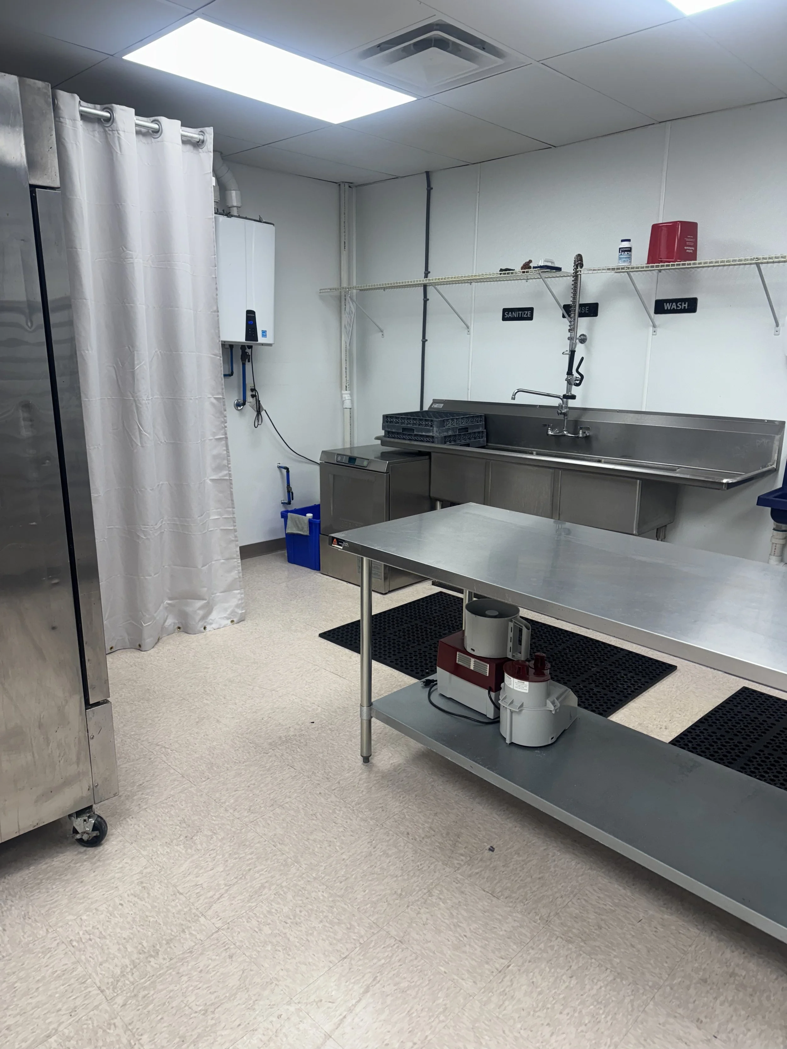 Commercial sanitized dishwashing area with stainless steel sink, work table, cleaning equipment, and labeled shelves for sanitizing, washing, and rinsing.