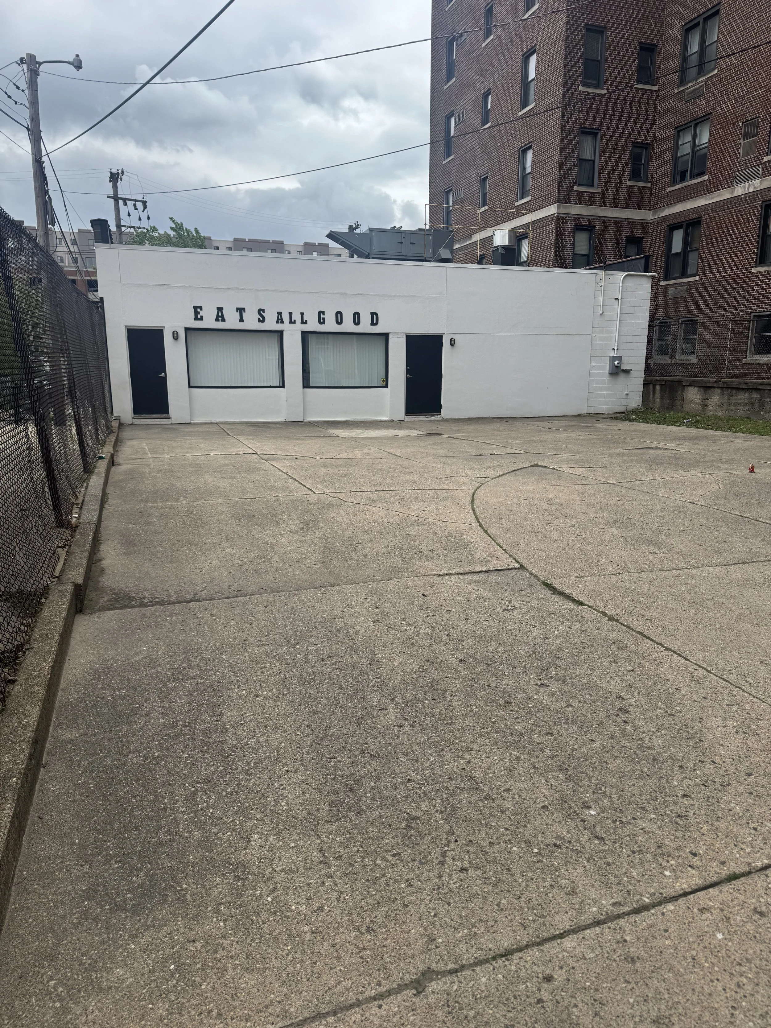 An outdoor concrete area with a white building at the back displaying the text 'EATS ALL GOOD' in black letters. The building has two black doors and two large windows, with an overcast sky above and multi-story brick buildings nearby.