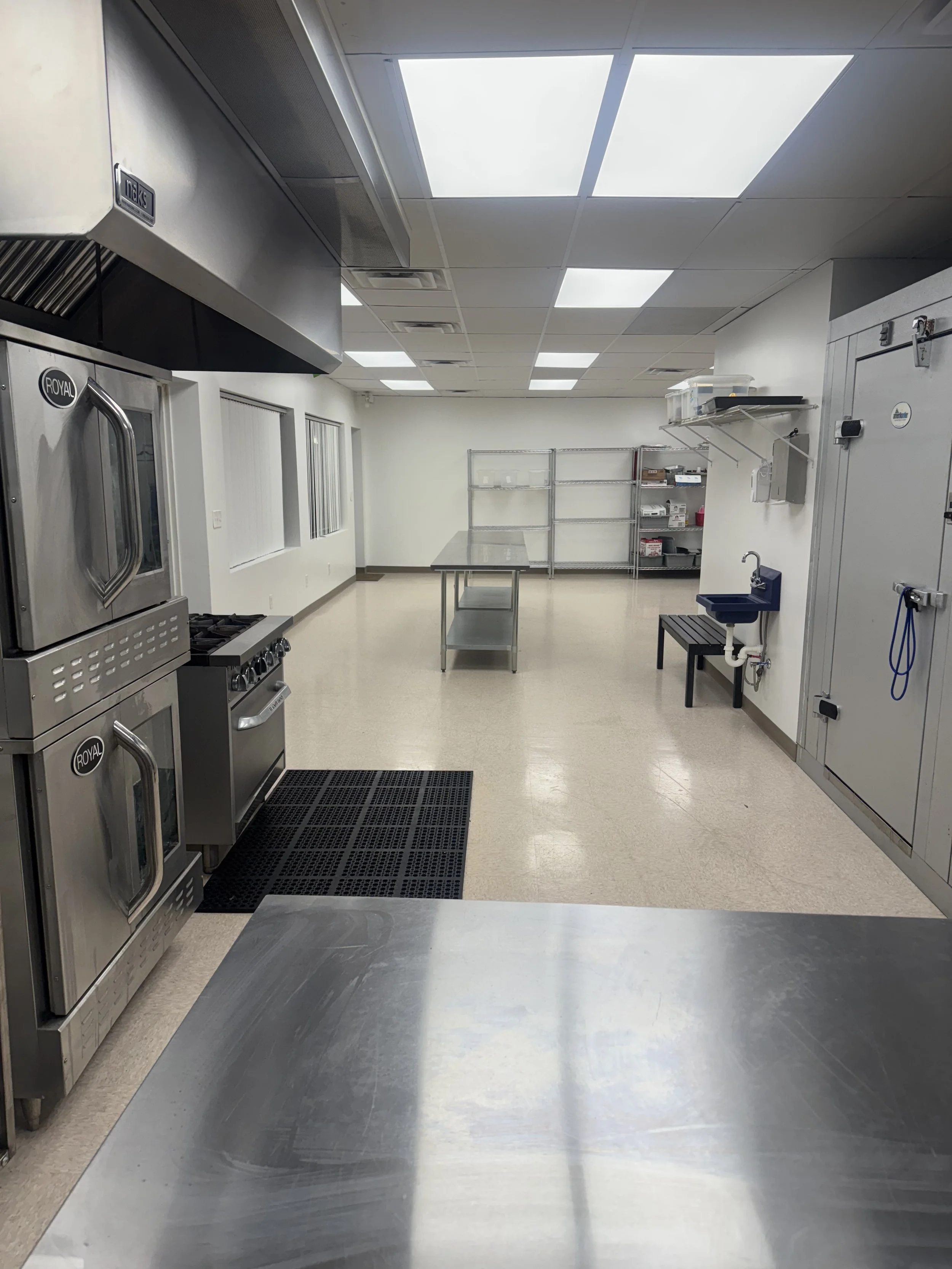 Commercial kitchen with stainless steel appliances, shelving units, a small sink, and a window, illuminated by ceiling lights and natural light.