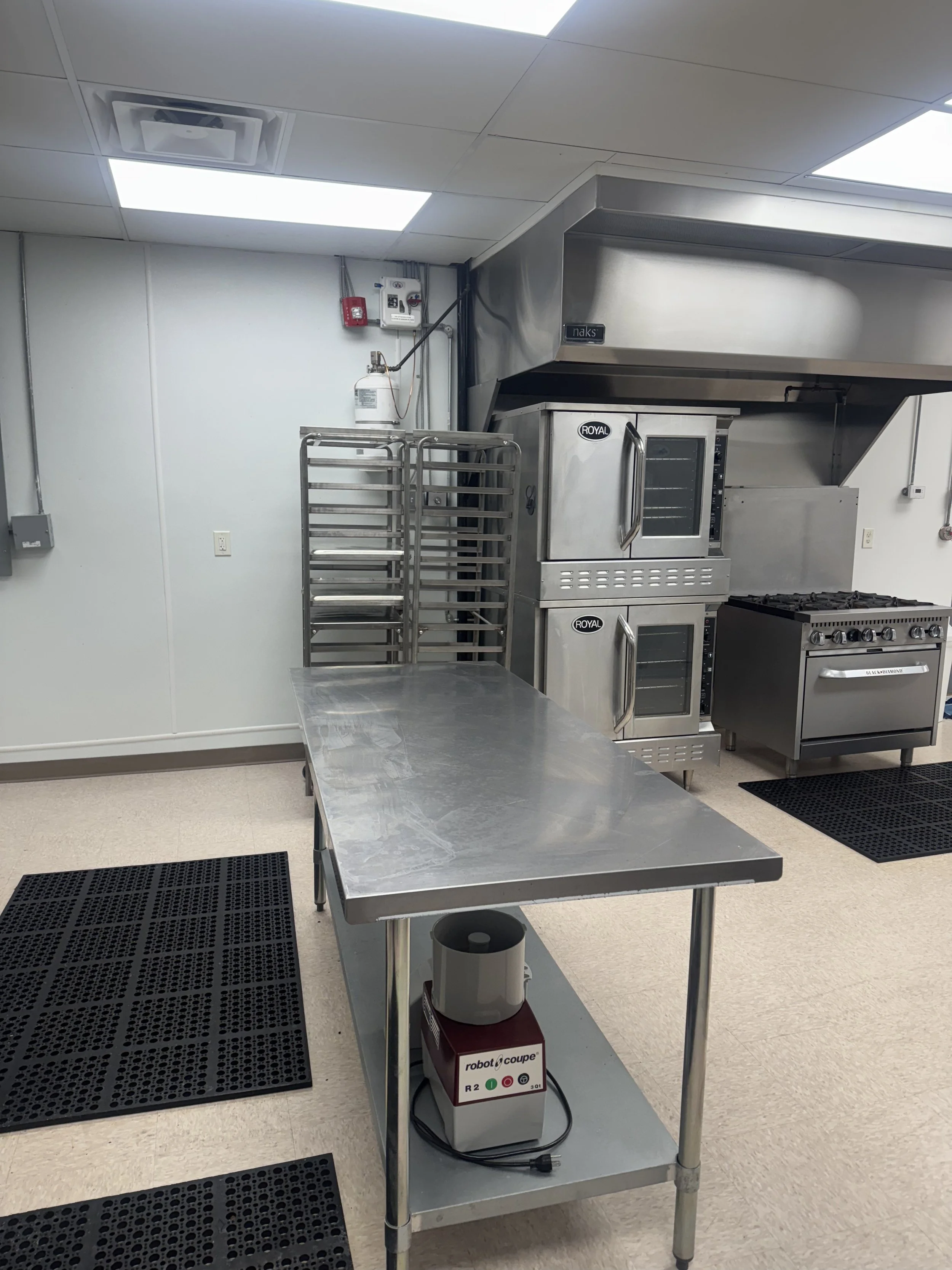 Commercial kitchen with stainless steel appliances, including ovens, a stove, a metal prep table, racks, and black rubber mats on the beige tiled floor.