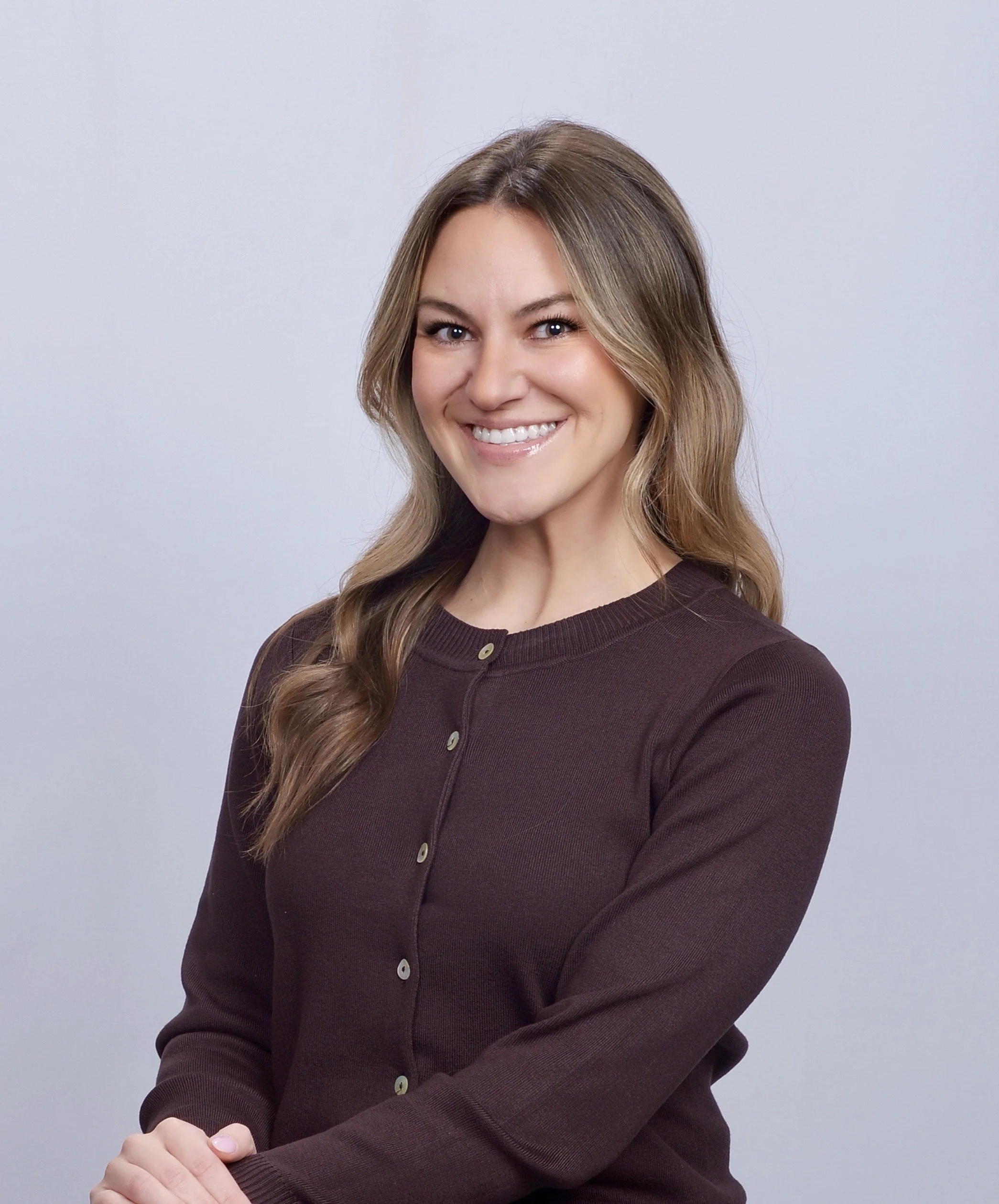 A smiling woman with long, wavy blonde hair wearing a dark purple button-up sweater, standing against a plain white background.