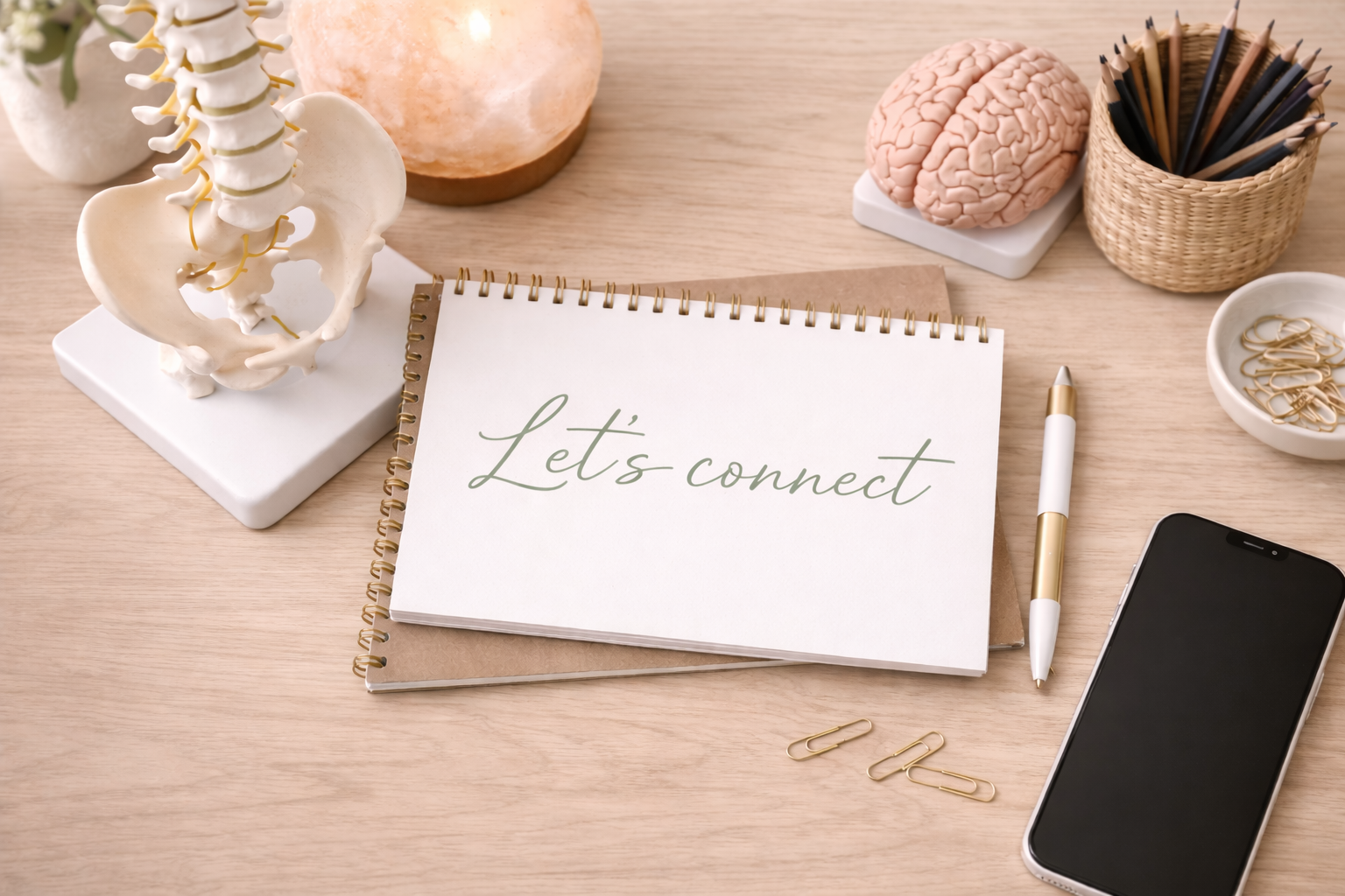 A wooden desk with a model of a human spine, a salt lamp, a salt lamp, a brain model, a basket of colored pencils, a dish of paper clips, notebooks, a pen, and a smartphone. The notebooks have a sign that reads 'Let's connect'.