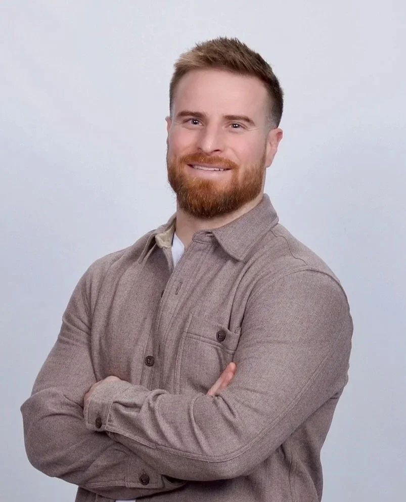 A young man with a beard and short hair, smiling, wearing a gray button-up shirt, standing with arms crossed against a plain light background.