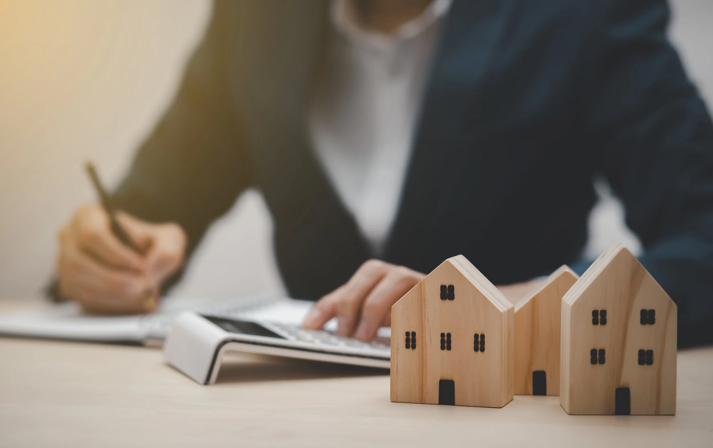 Person in a dark blazer and white shirt sitting at a desk, using a calculator and writing in a notebook, with three wooden house-shaped models with black windows and doors in the foreground.