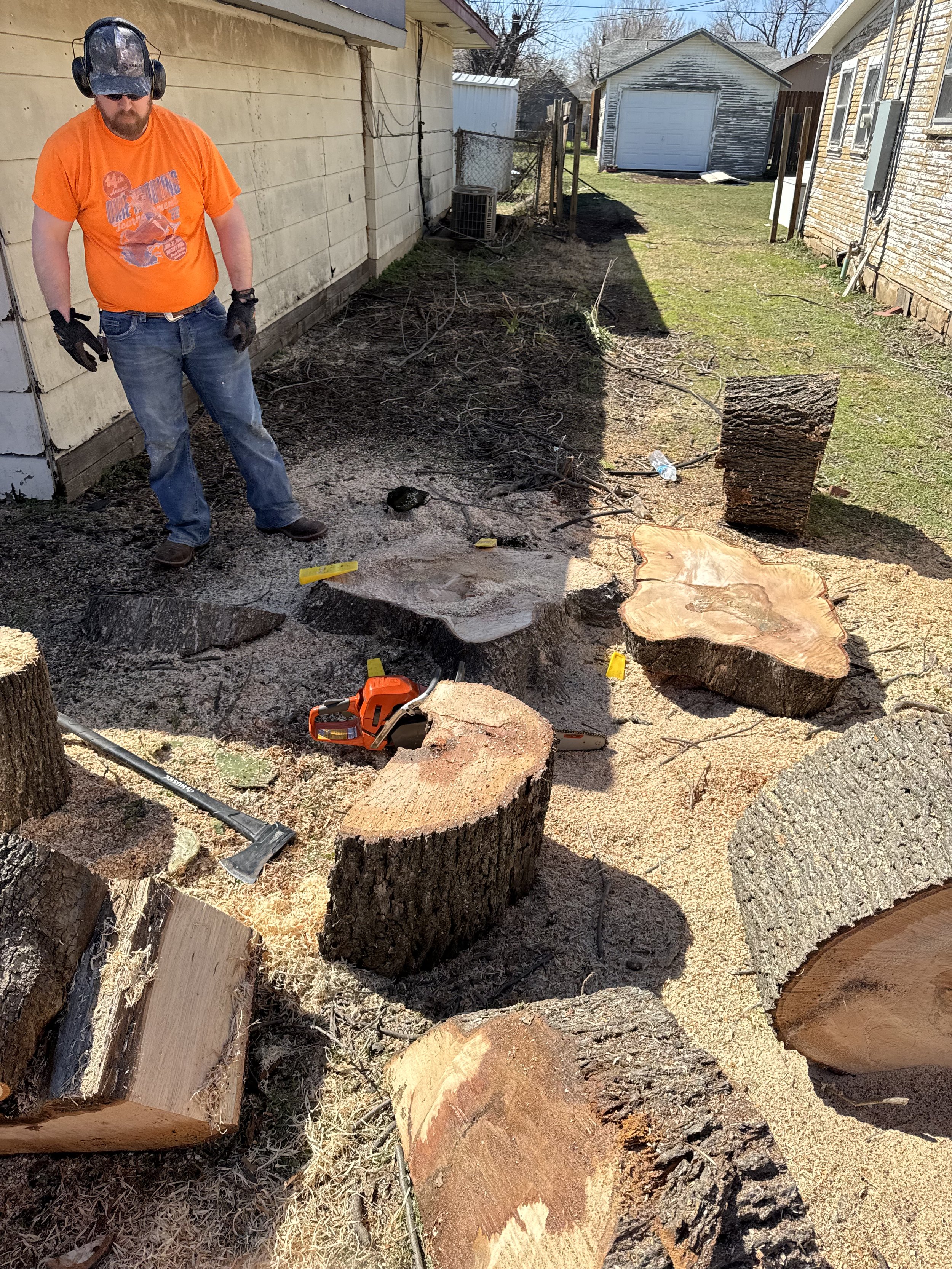 A man in orange shirt, jeans, and safety gear is standing in a backyard with cut tree logs, a chainsaw, and wood debris, preparing to cut or haul the wood.
