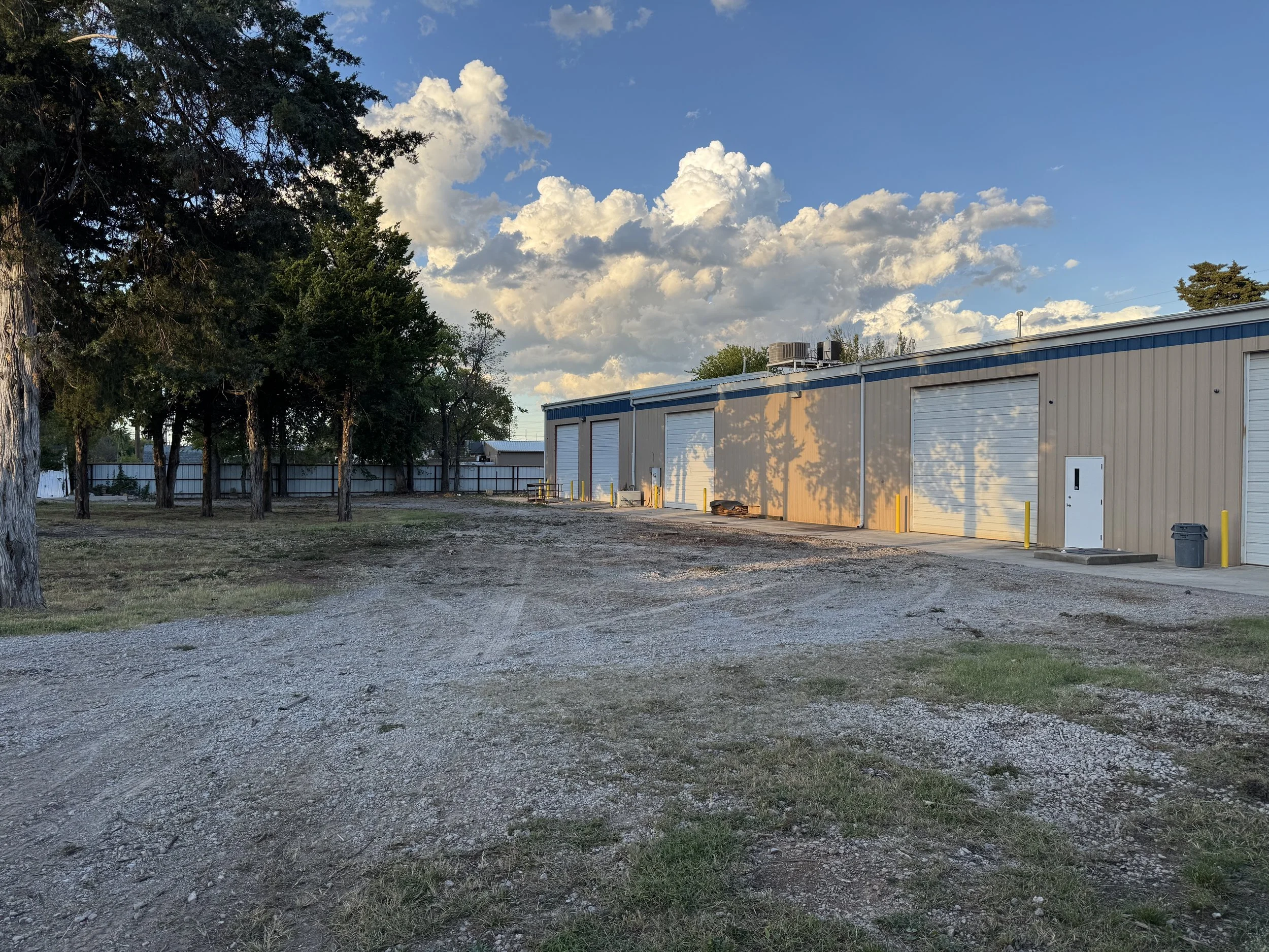 Sunset over a warehouse with several large garage doors, surrounded by trees and a gravel driveway.