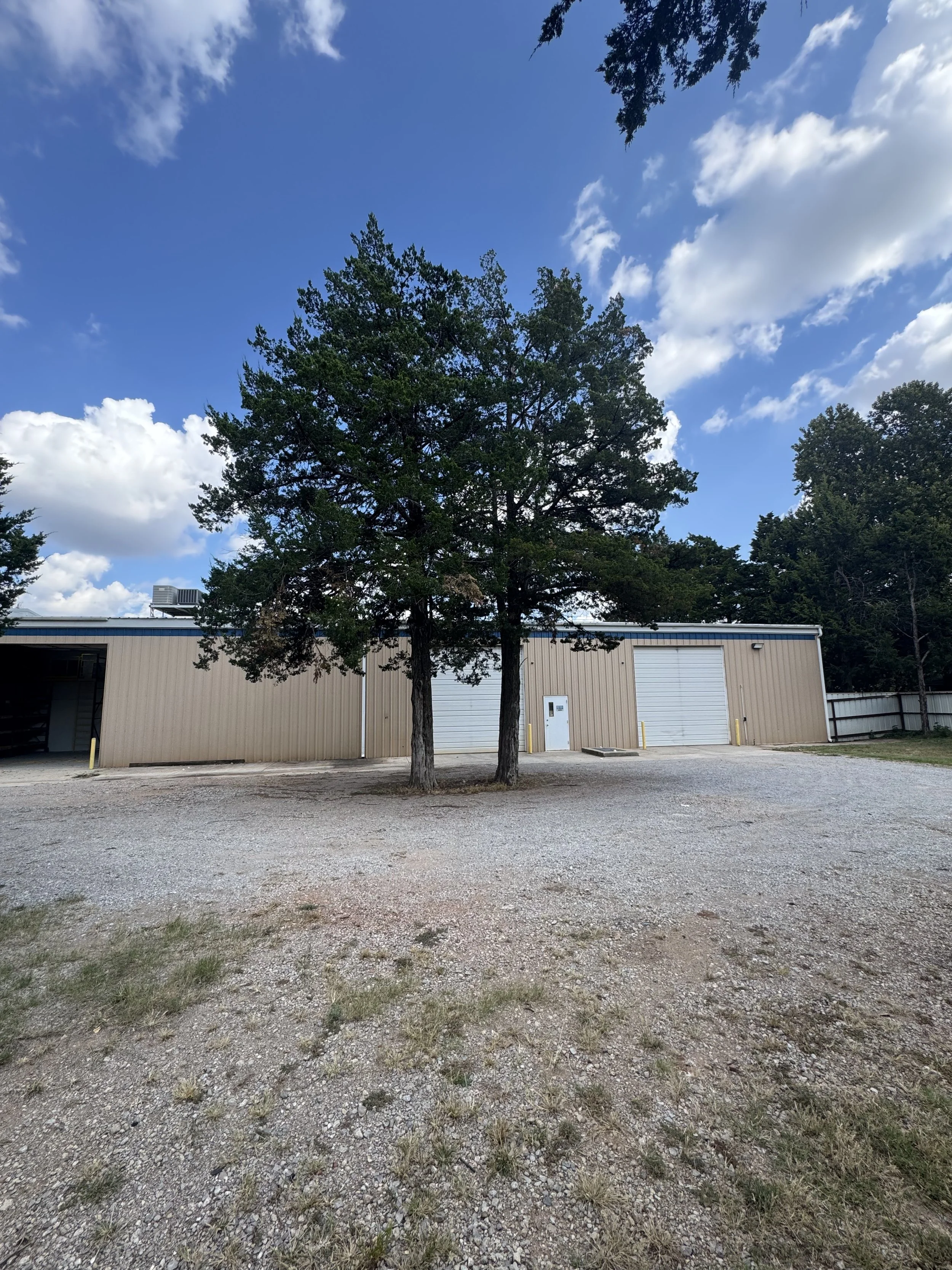 Two trees in front of a beige industrial building with two garage doors, a small side door, and a gravel parking area under a partly cloudy sky.