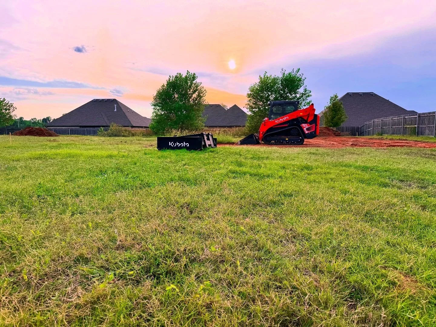 A grassy backyard with a Kubota mini excavator and an overturned black Kubota container, with some dirt mounds and houses in the background during sunset.