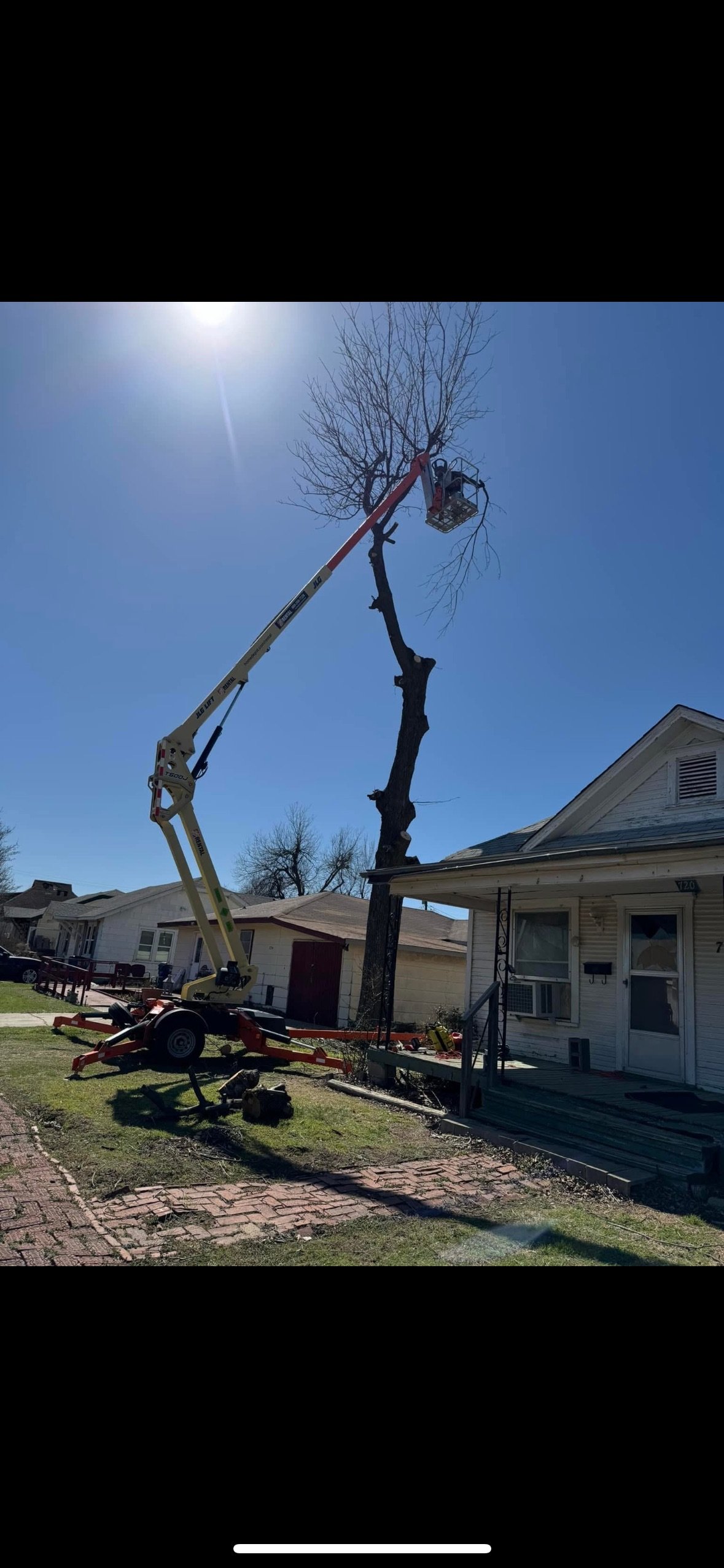 A worker in a cherry picker trimming a tall, leafless tree in front of a house on a sunny day.