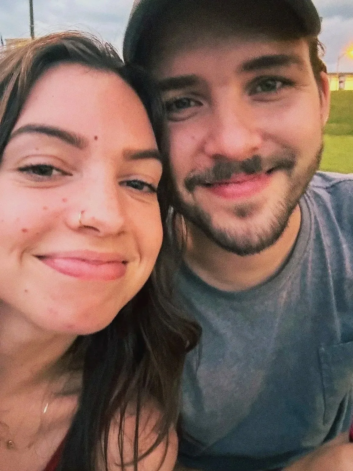 Close-up of a young couple smiling, outdoors during the evening. The woman has long dark hair, wears a nose ring, and is smiling. The man has short dark hair and a beard, with a slight smile. Background shows a grassy area and some lights.