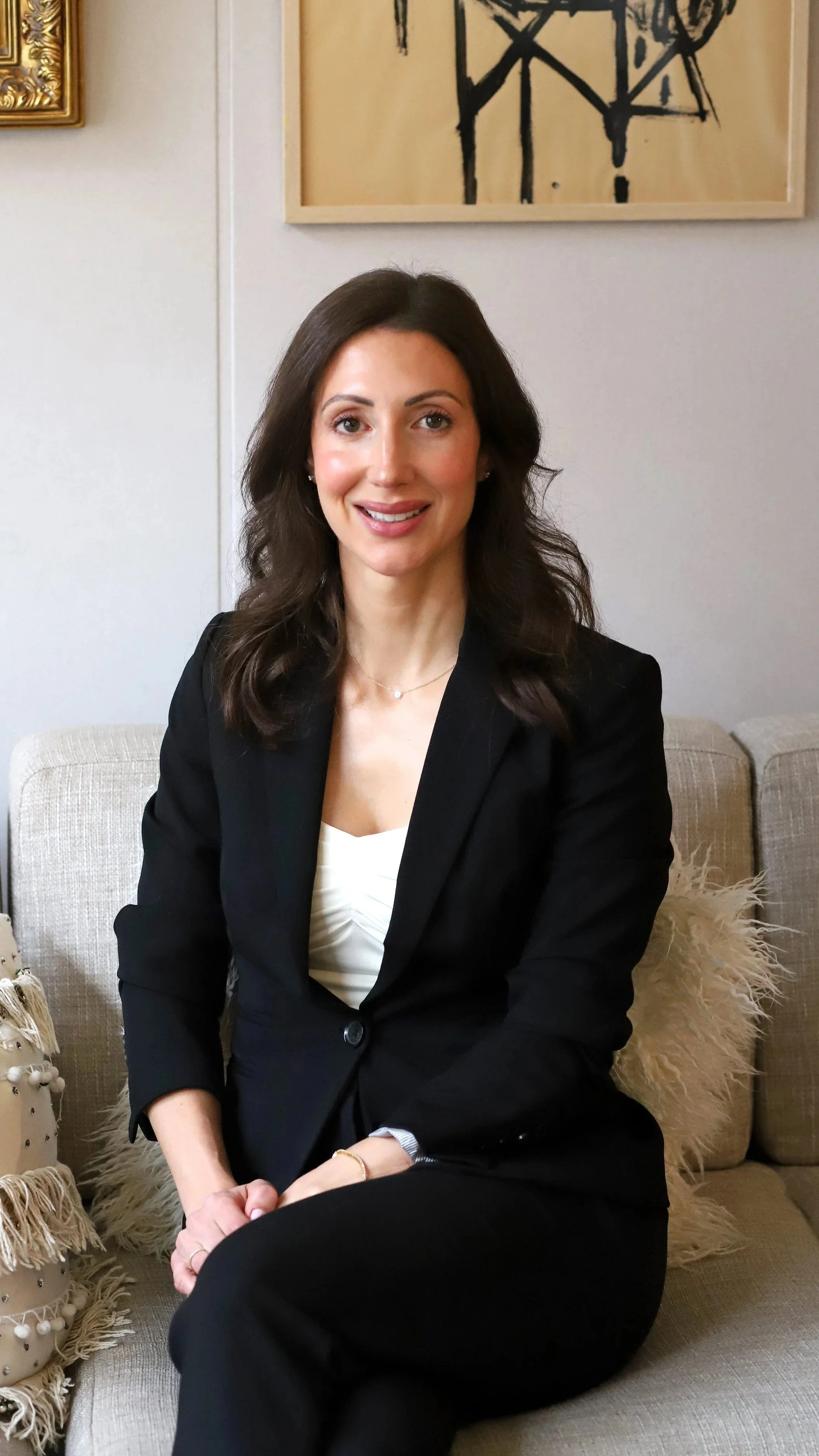 A woman sitting on a beige sofa in a black blazer and white top, smiling at the camera. Behind her are framed artworks on a white wall.