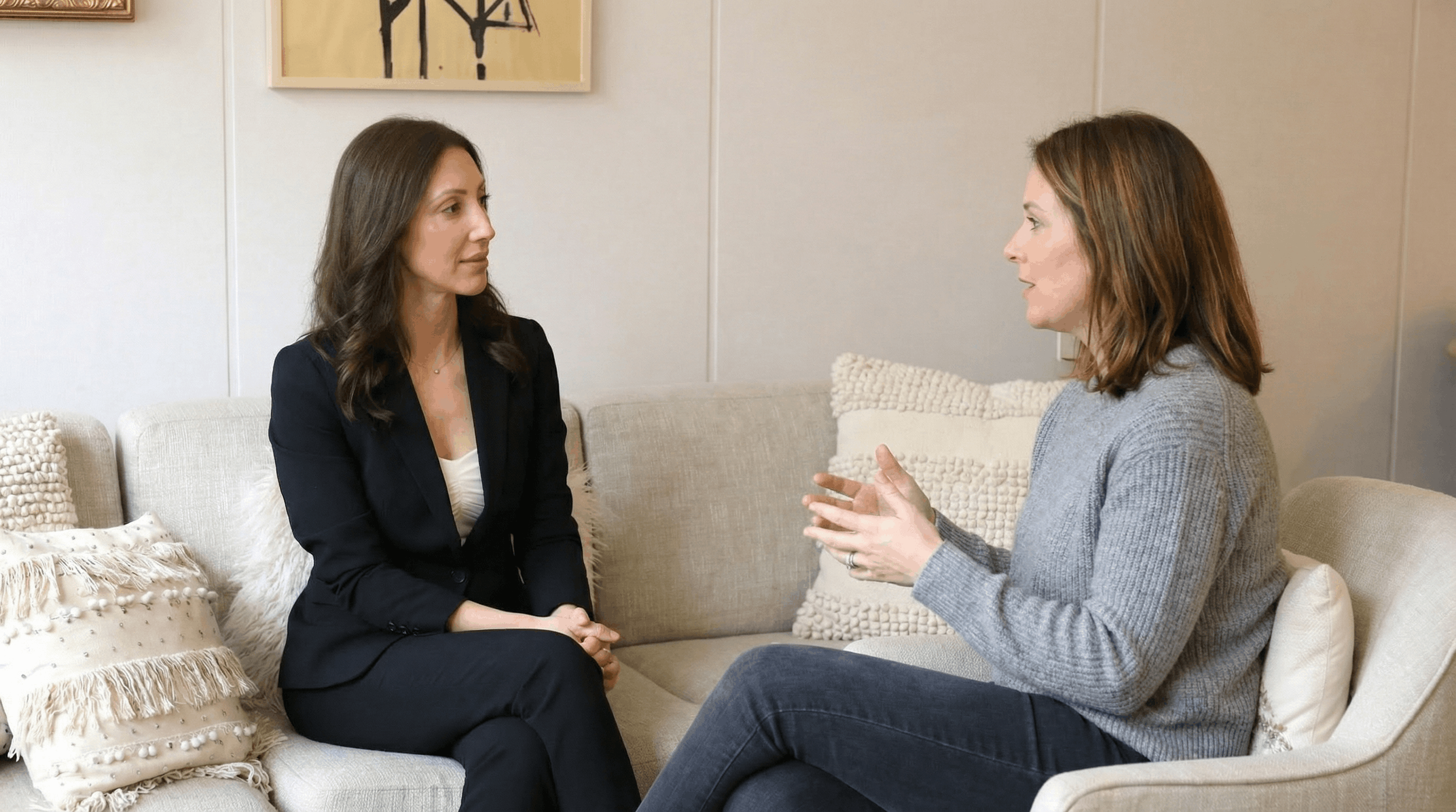 Two women sitting on a beige sofa, engaged in conversation in a modern room with neutral decor and framed artwork on the wall.