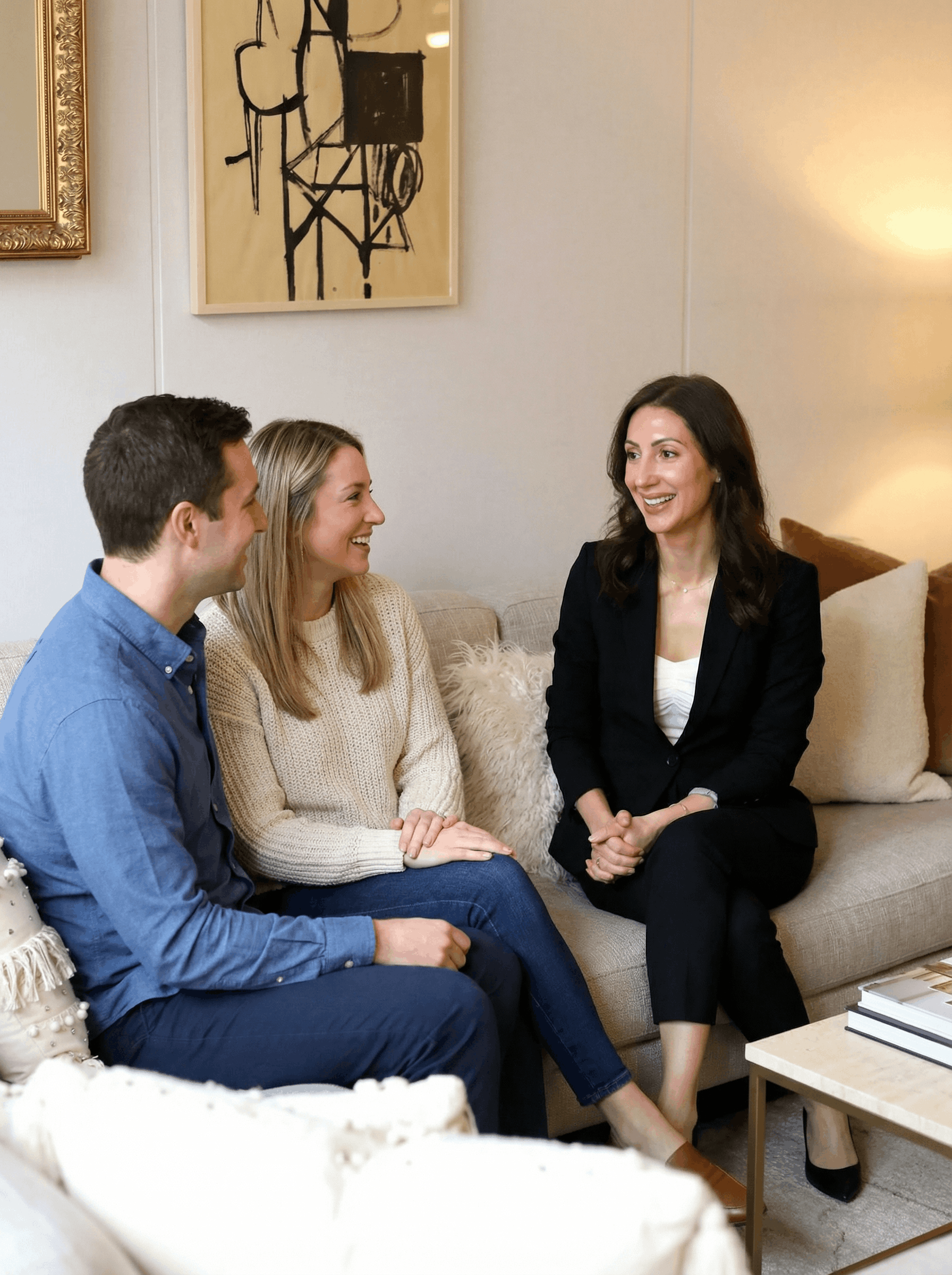 A woman in a black suit is sitting on a beige couch with two young people, a man and a woman, in casual clothing. They are smiling and appear to be having a friendly conversation in a cozy living room.