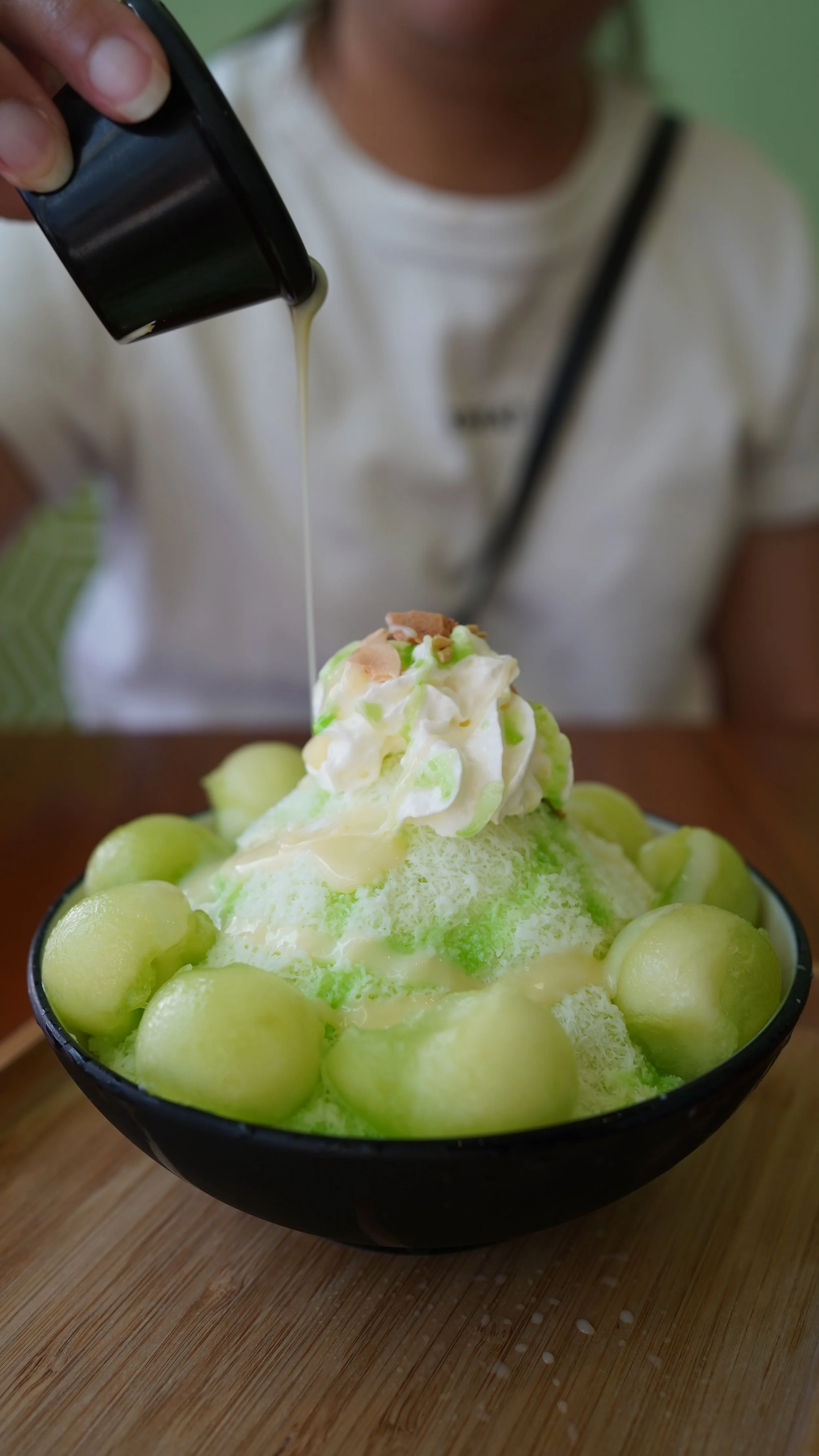 A bowl of honeydew melon Korean bingsu shaved ice topped with whipped cream, nuts, and green syrup with a person pouring condensed milk over it.