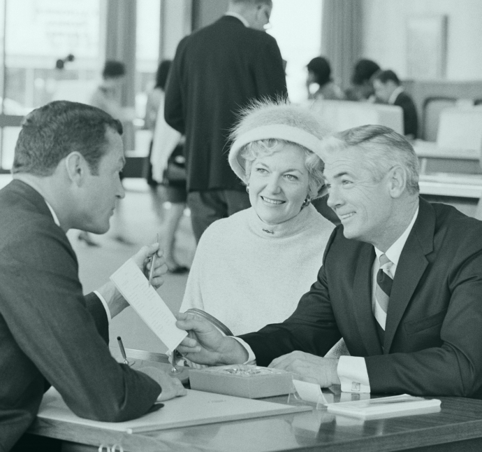 A couple is seated at a desk, smiling and talking to a man in a suit, while a woman in a white hat with a feather looks on, in an office setting.
