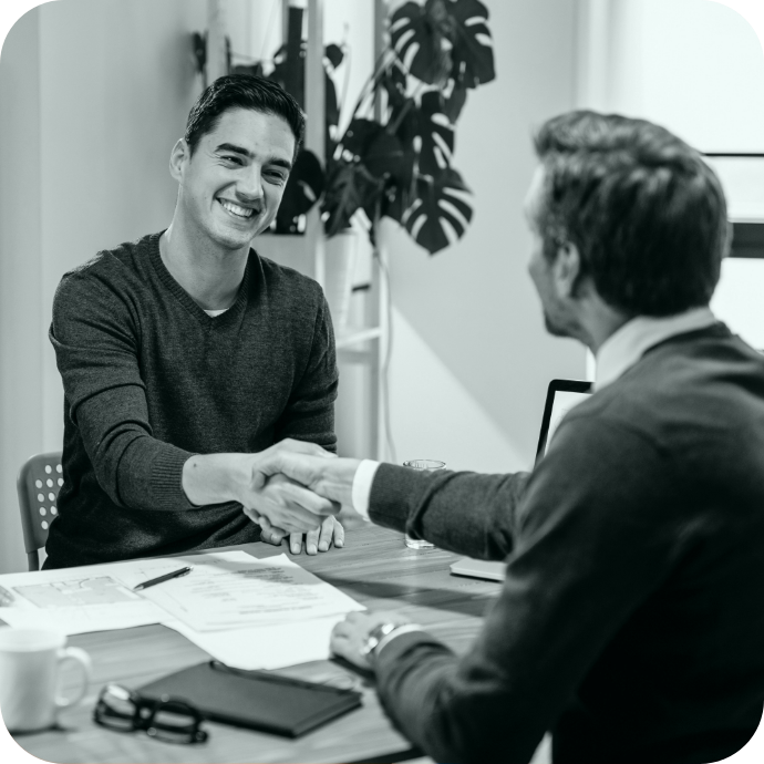 Two men shaking hands during a business meeting in a modern office, with documents, a laptop, and a coffee mug on the table.