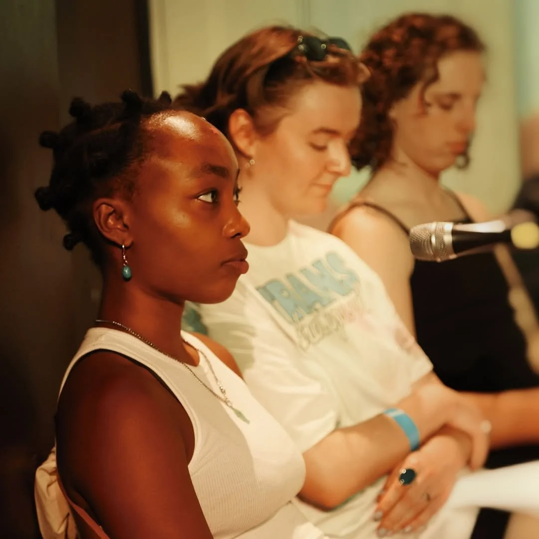 Three women sitting at a table in a row, with a microphone in front of the woman on the right. The woman in the foreground is African American, with her hair styled in bantu knots, wearing a white sleeveless top, earrings, and a necklace, looking attentively to her left. The middle woman has red hair, wearing a graphic t-shirt, with a gentle expression. The third woman has curly hair, wearing a dark sleeveless top, with her eyes closed or looking down.
