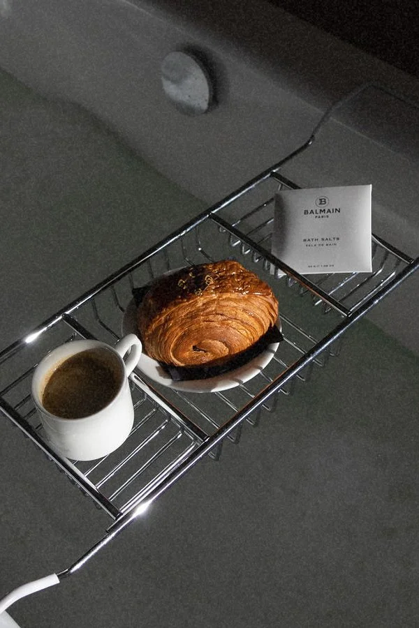 A breakfast tray with a croissant on a plate, a cup of coffee, and a packet of Bath Salts on a metal rack.