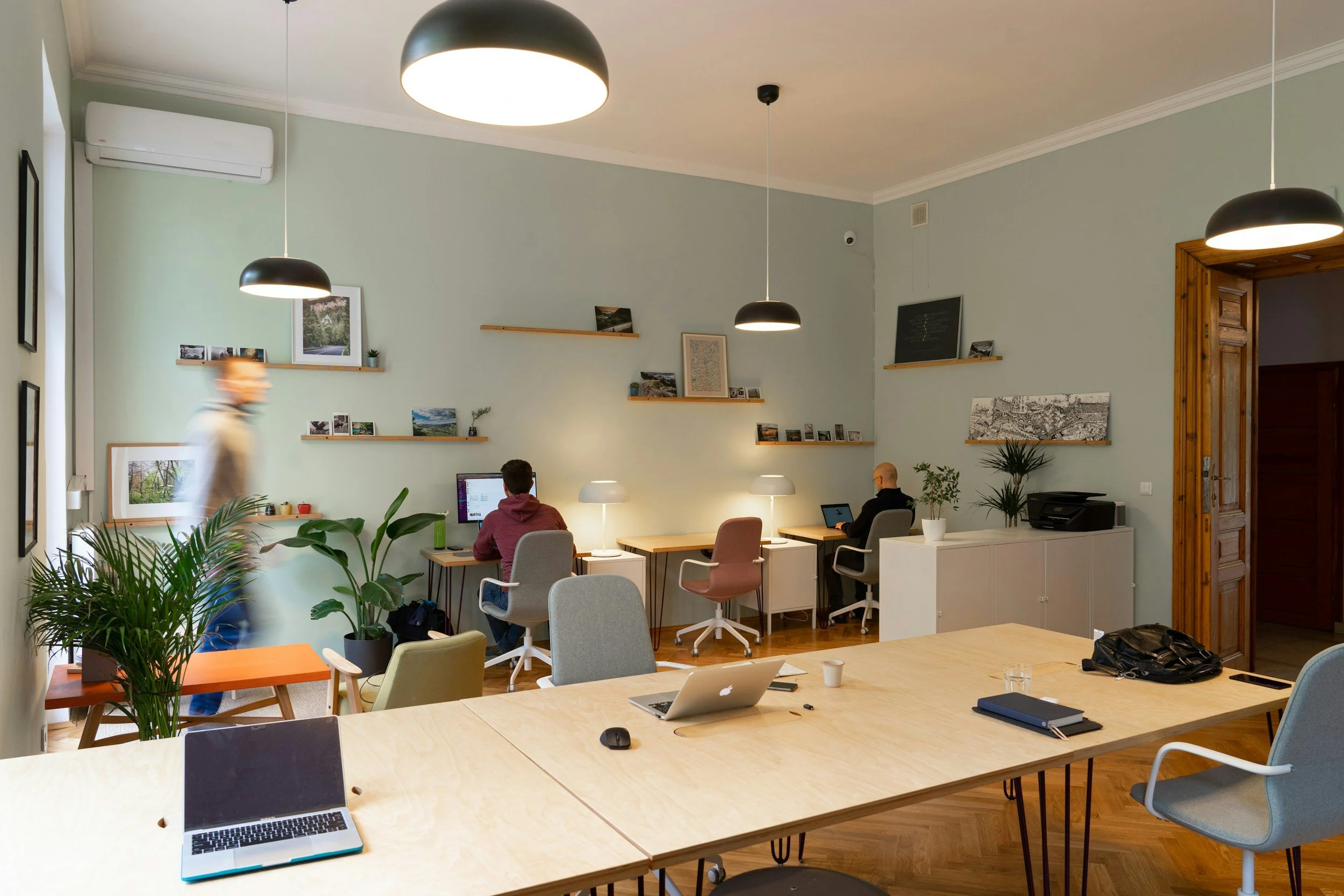 Modern office workspace with three people working at desks against a light green wall, featuring shelves with pictures and decor, green plants, laptops, notebooks, and a conference table in the foreground.
