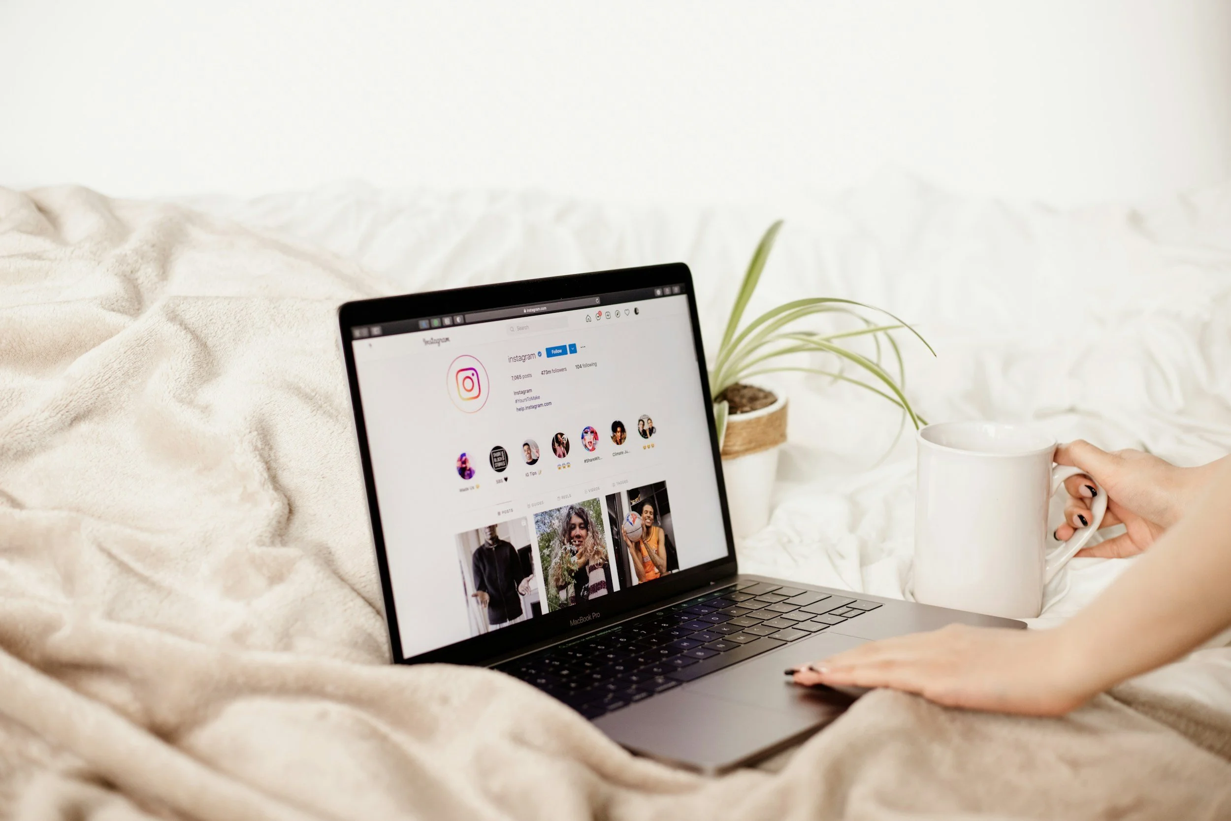 A person with black nail polish holding a white coffee mug next to a MacBook Pro laptop on a bed with beige blankets. The laptop screen displays an Instagram profile page, and there is a potted plant nearby.