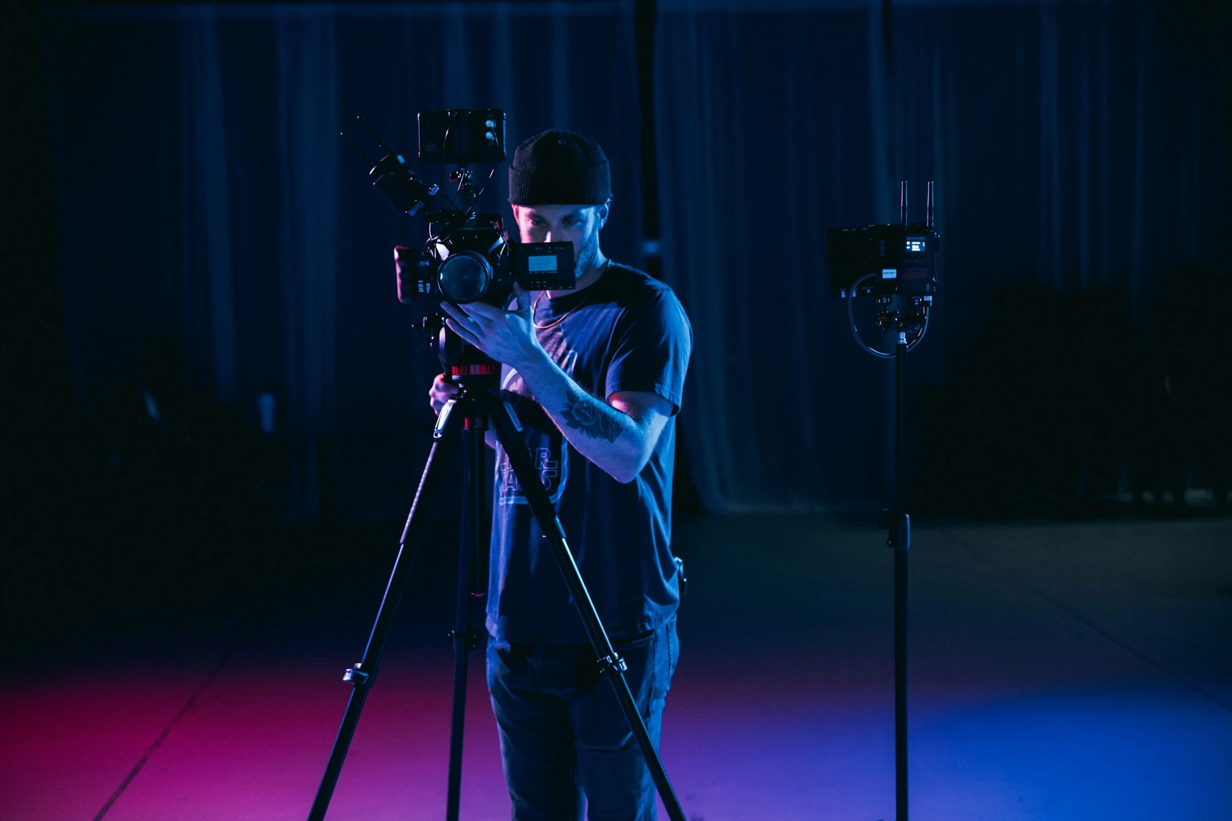 A person wearing a black beanie operating a professional camera on a tripod in a dark room with blue and purple lighting, with additional camera equipment on a stand nearby.
