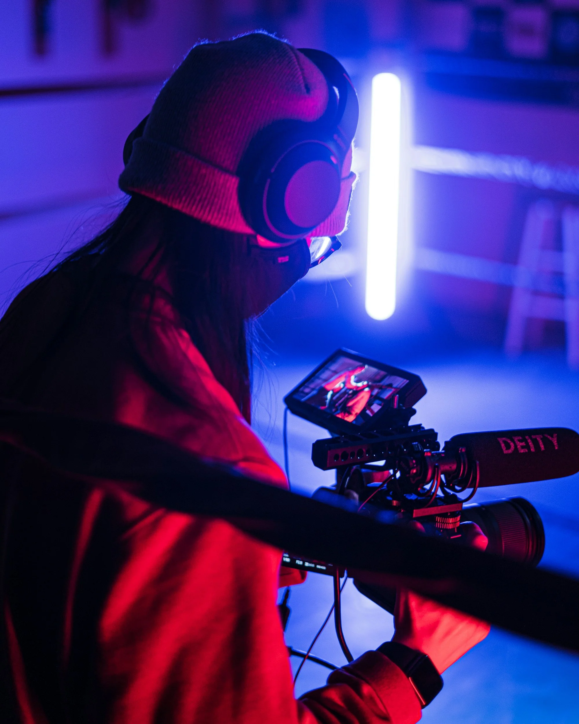 A person in a gray beanie and red hoodie filming with a professional camera and microphone in a dimly lit room with purple and blue neon lighting.